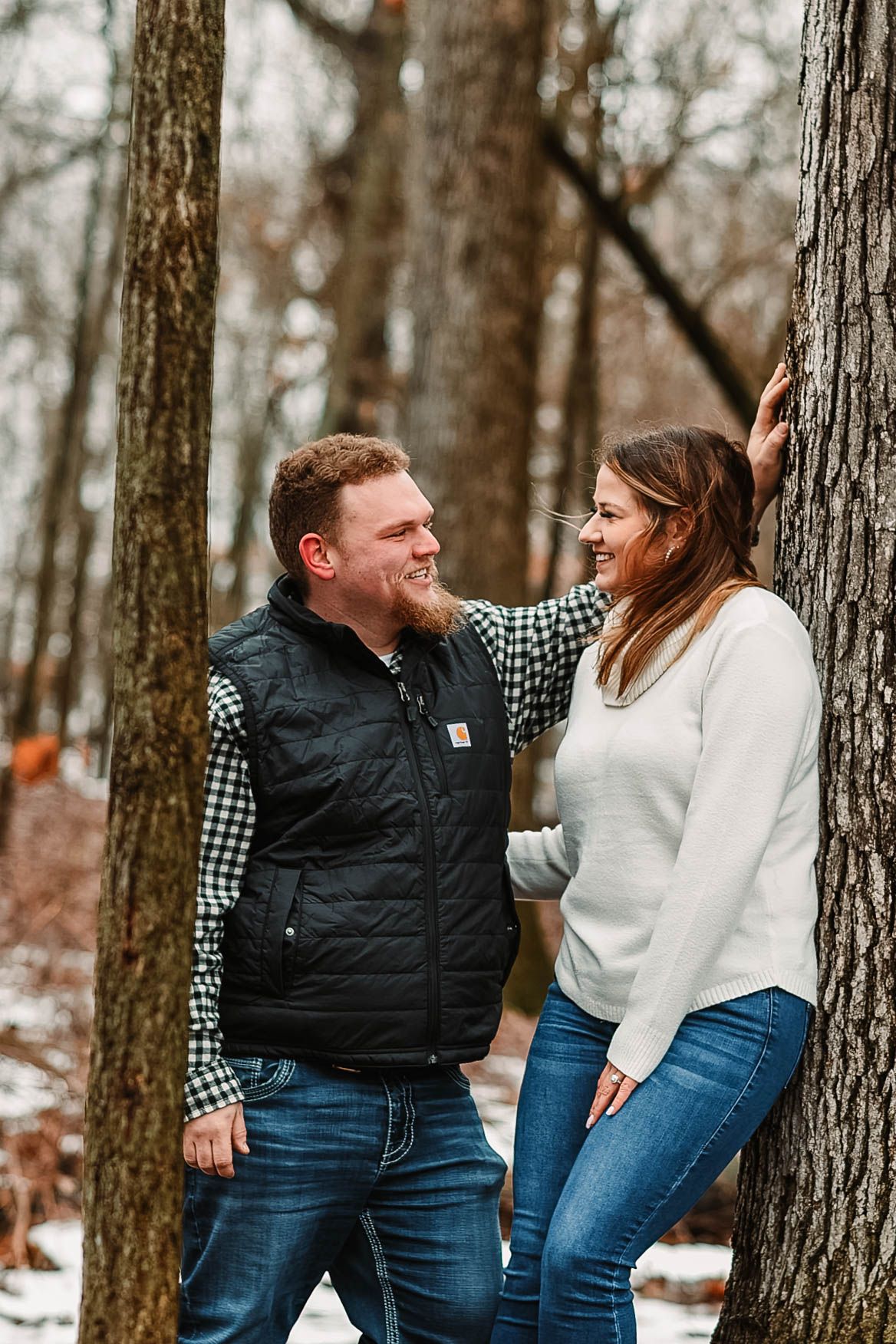A couple shares a playful moment between trees in a winter forest setting.