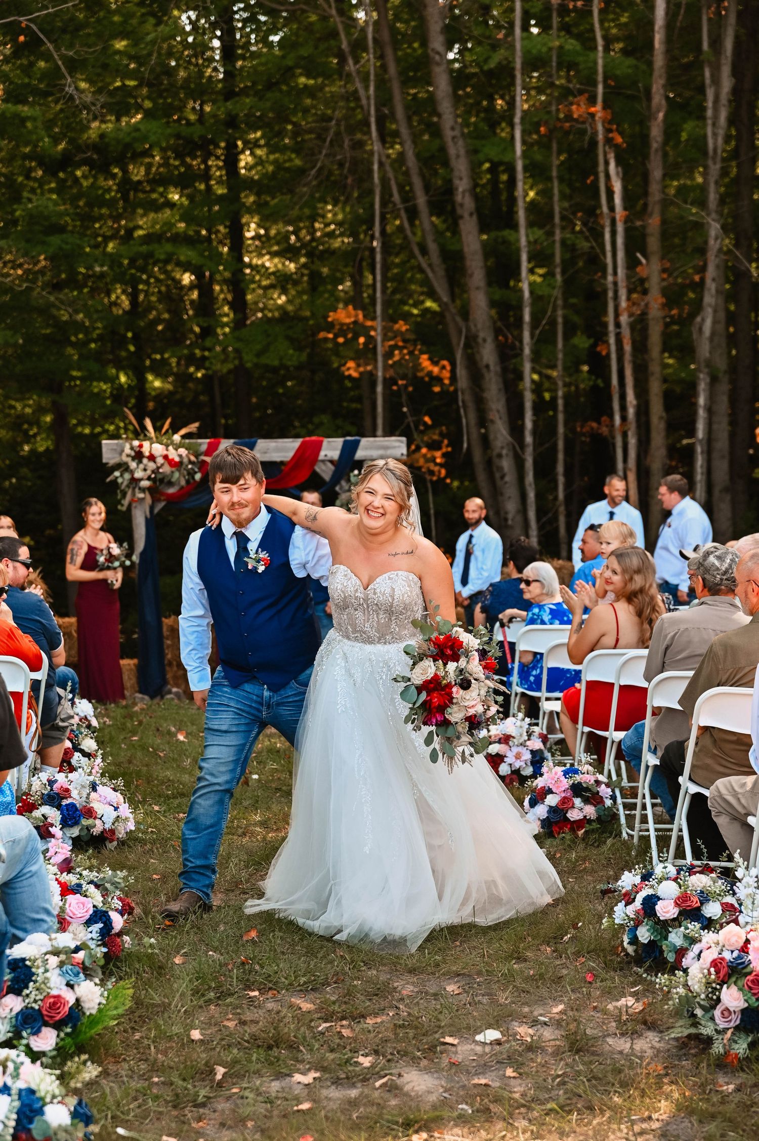 Outdoor wedding ceremony setup in a wooded area with white chairs and floral decorations.