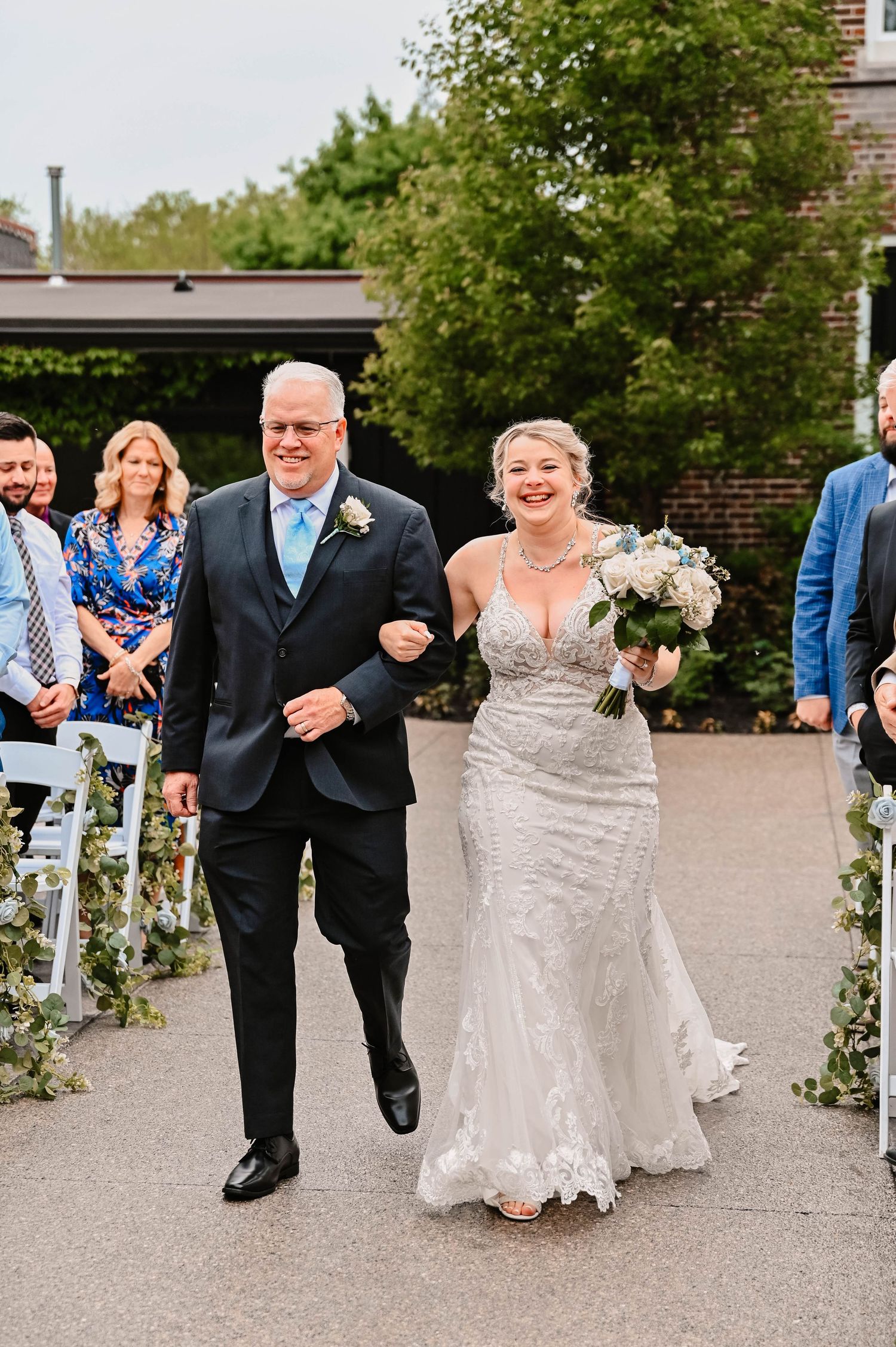 Wedding party members skip joyfully down an outdoor aisle lined with white chairs.