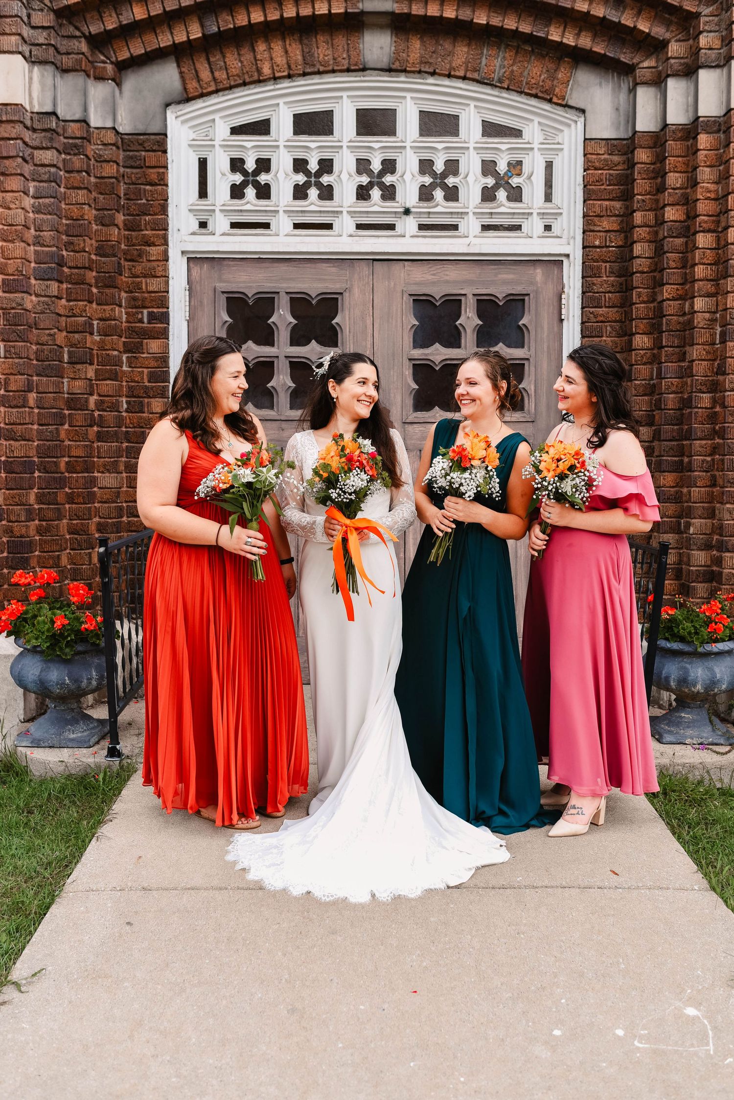 Wedding party in colorful dresses poses in front of ornate brick church entrance with flowers.
