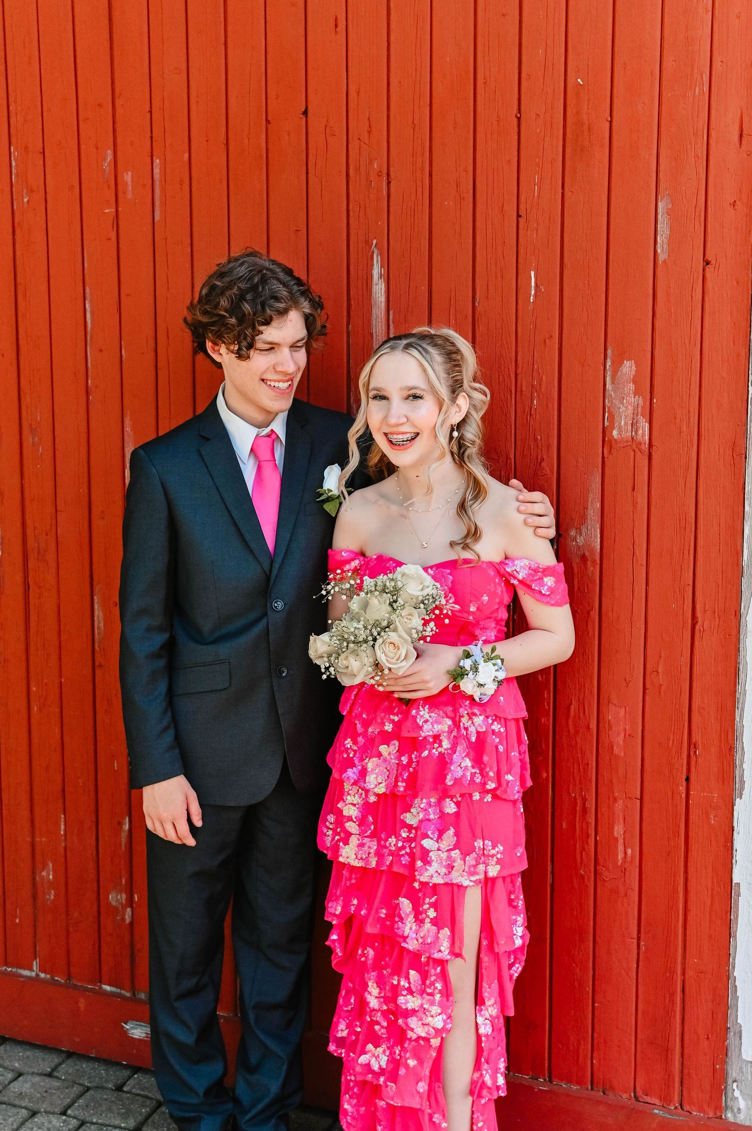 Couple in formal attire poses against a rustic red barn wall with matching pink dress and tie.