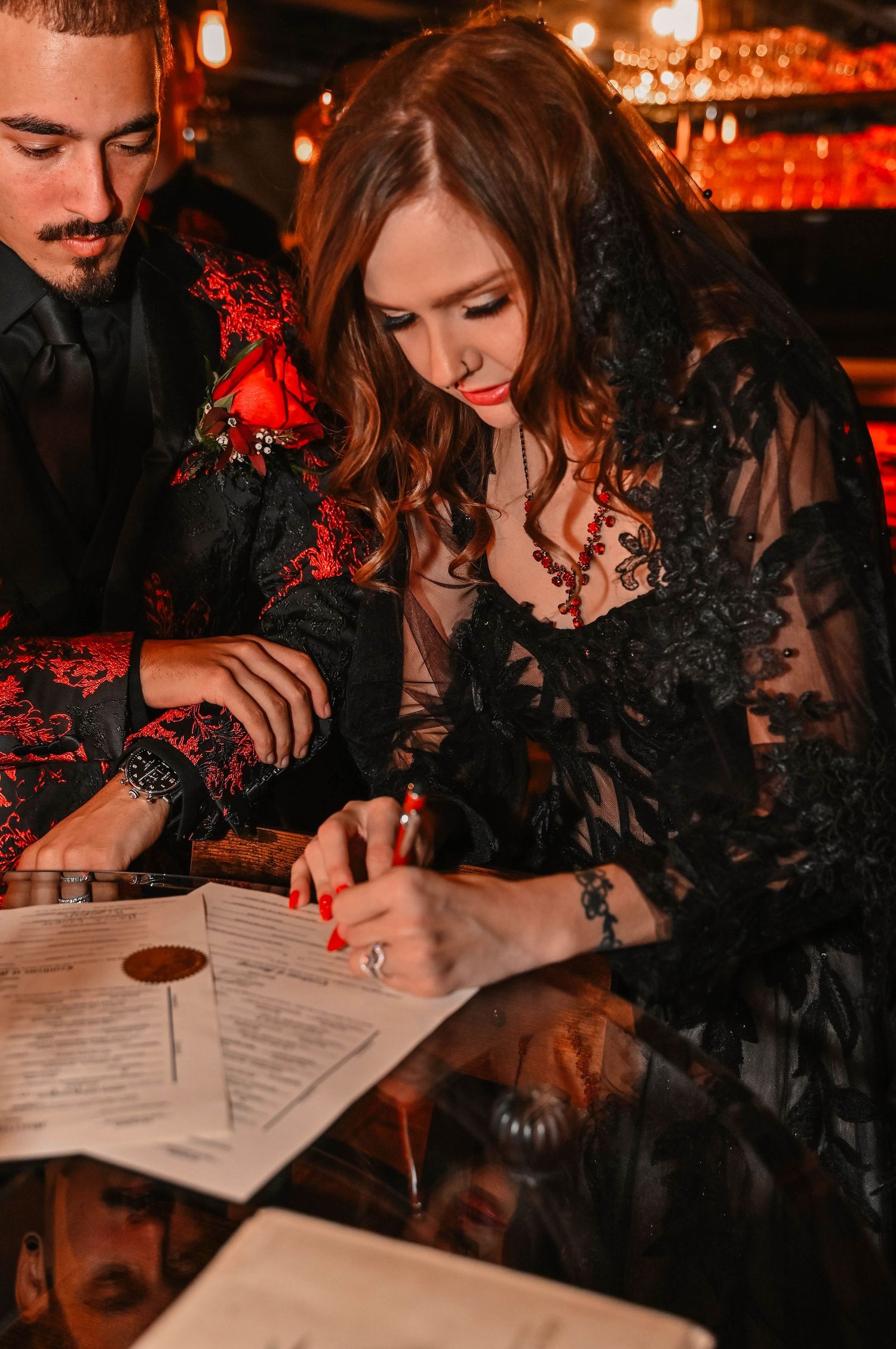 Person in black lace signing documents under moody red lighting.