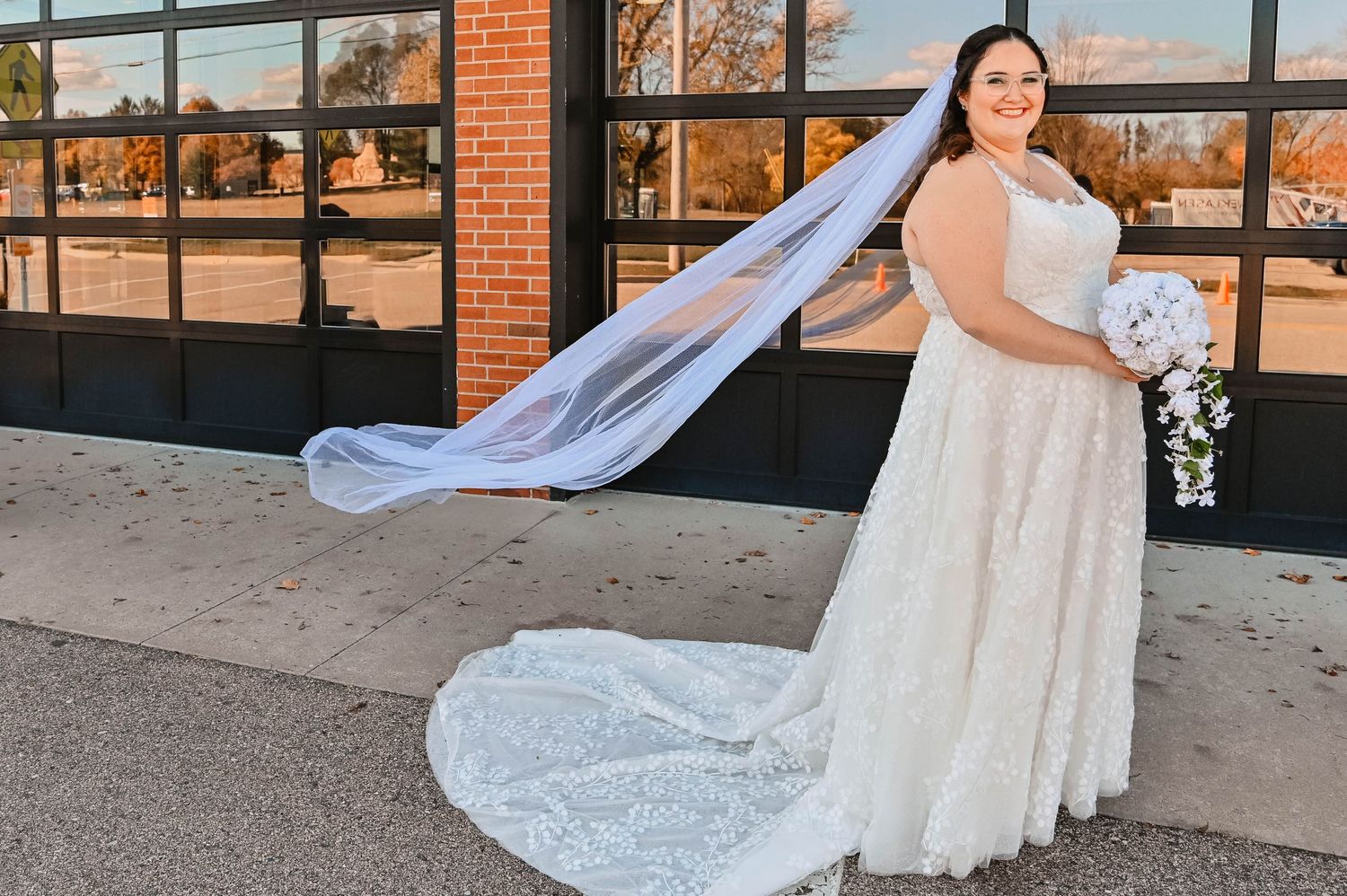 A long cathedral veil flows dramatically behind a bride in a lace gown against an industrial backdrop.