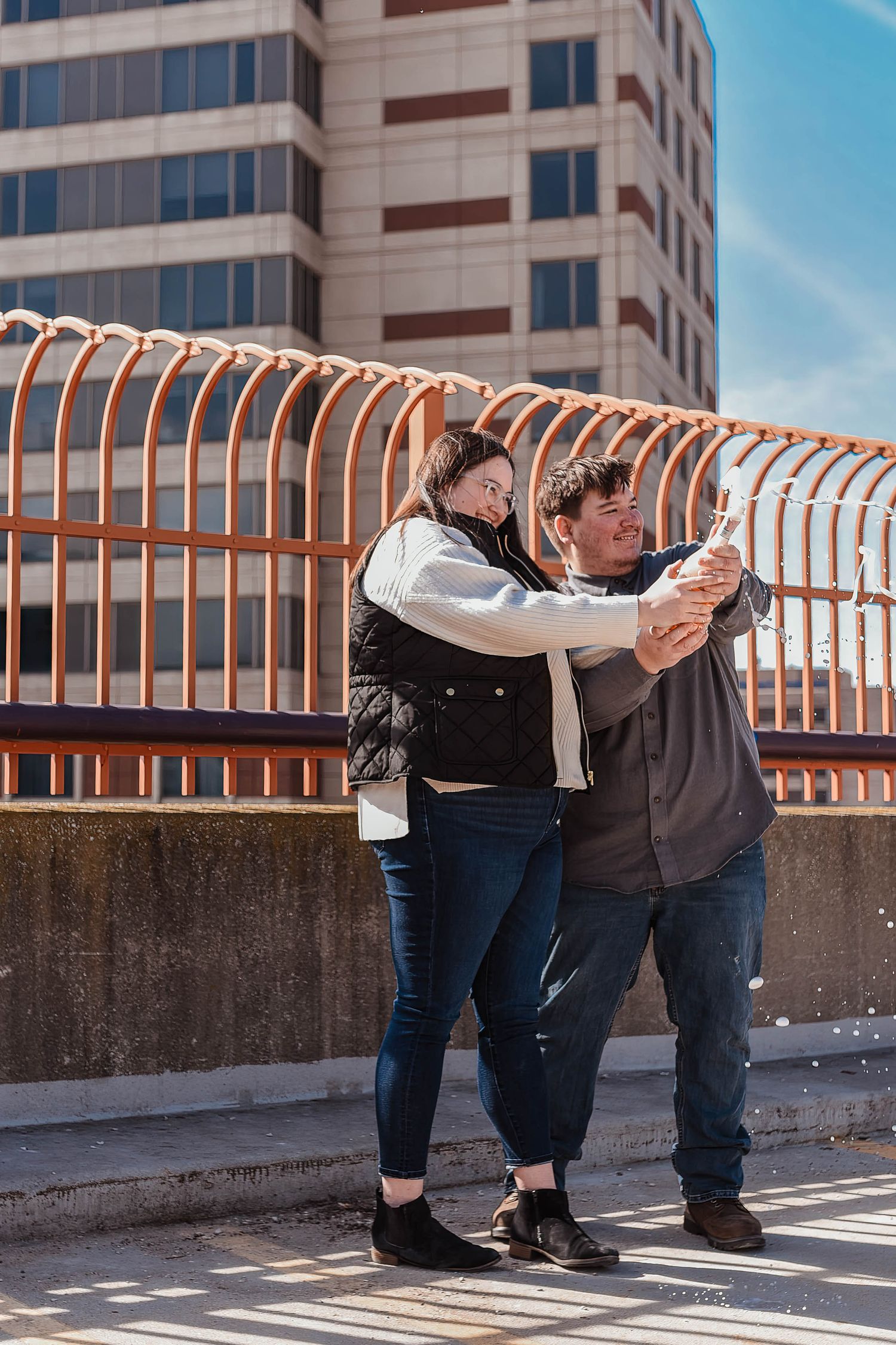 A romantic couple shares a playful moment against a railing with a city building in the background.
