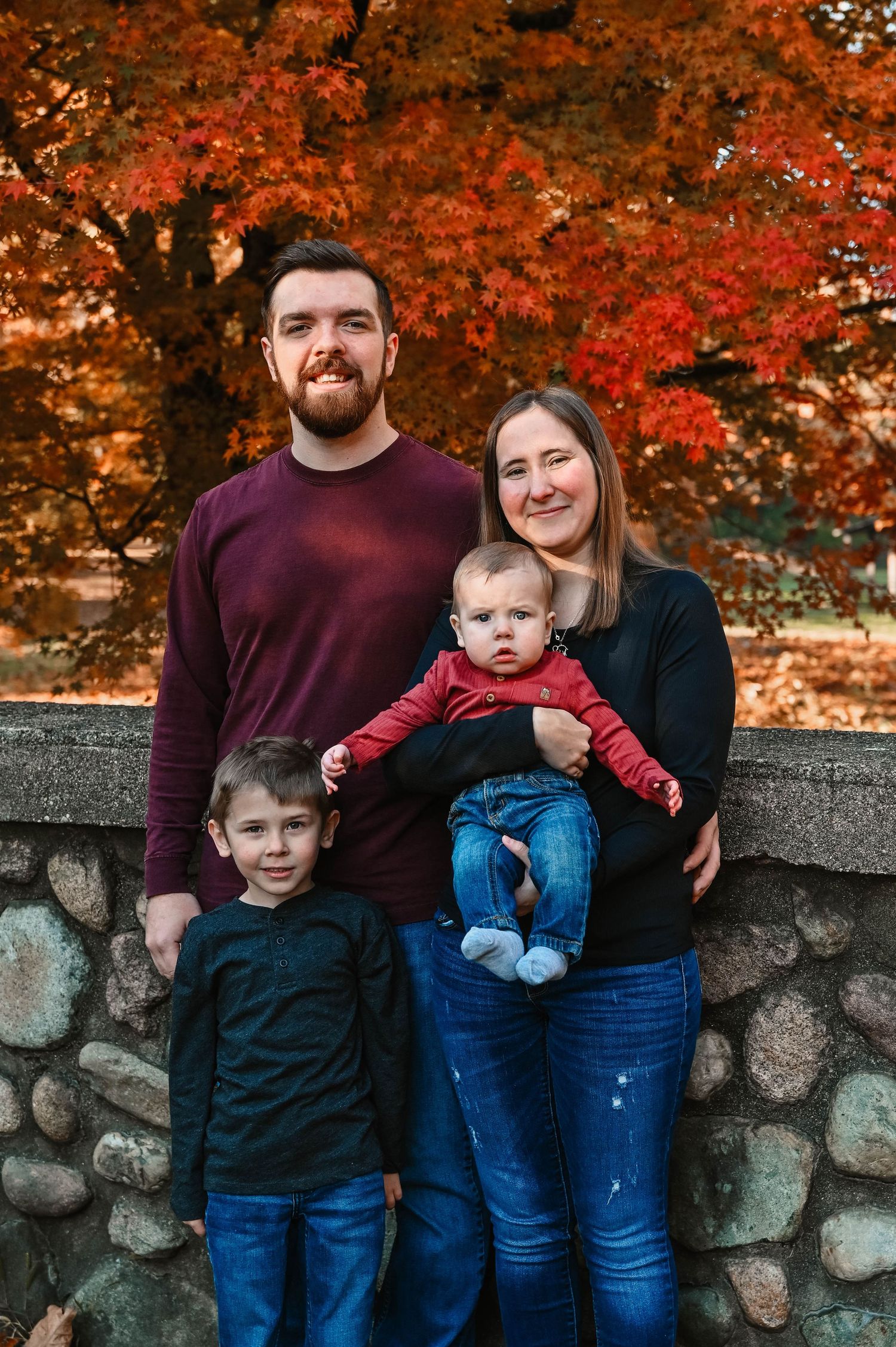 Family of four poses against a stone wall with vibrant red autumn foliage in the background.