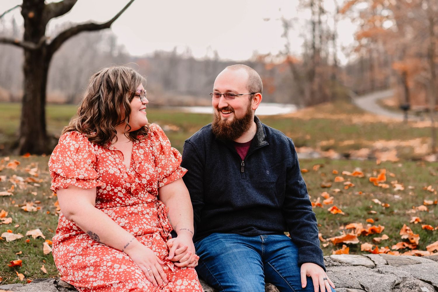 Couple in red dress and navy sweater share a joyful moment on fallen autumn leaves.