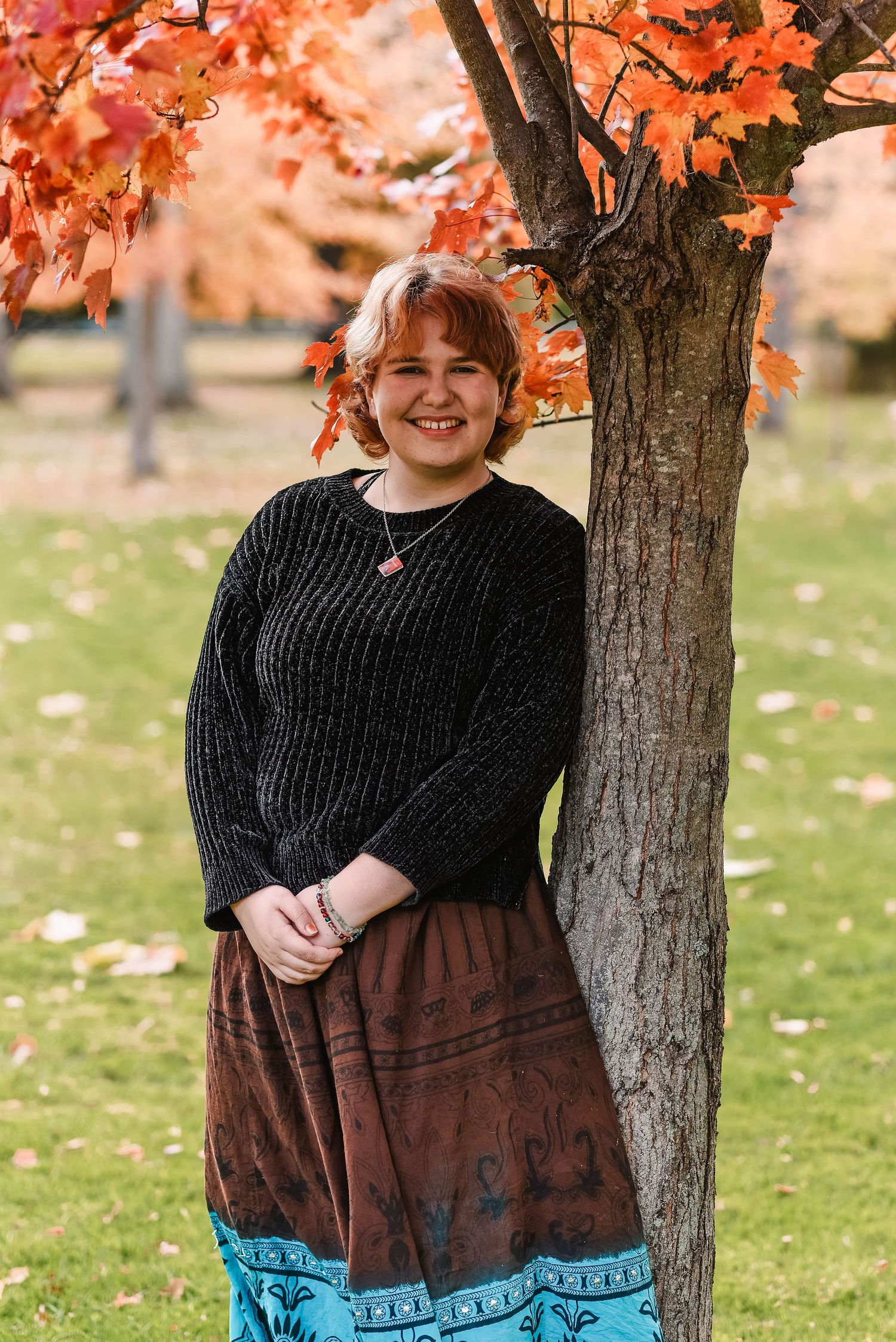 Person in black sweater and blue skirt poses under tree with orange autumn leaves.