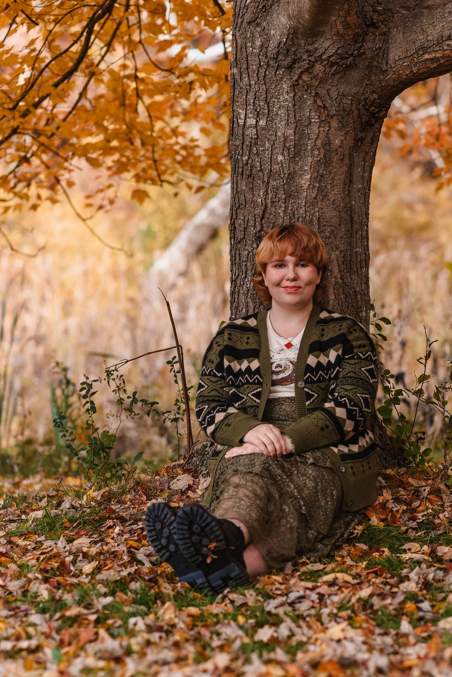 Autumn portrait session with patterned sweater sitting against large tree trunk.