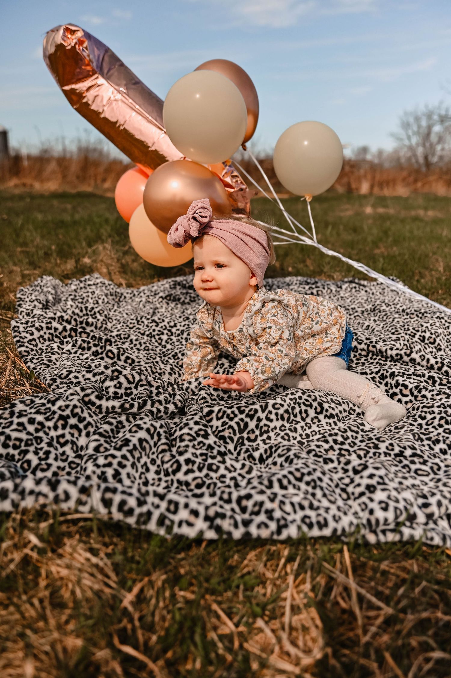 Infant plays with balloons while sitting on animal print blanket during outdoor photoshoot.