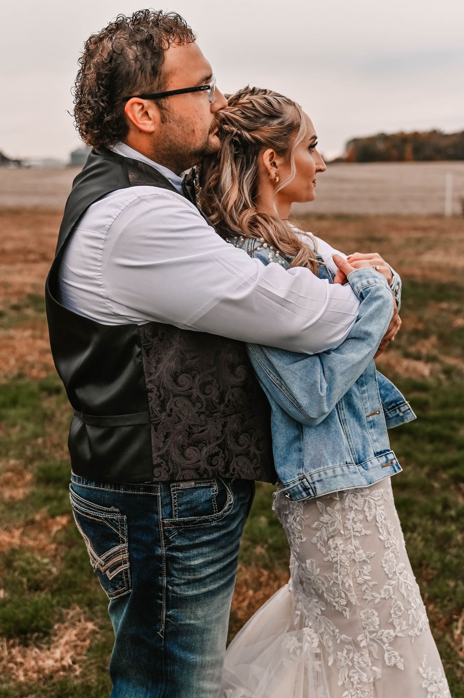 Couple embracing in casual denim and formal wear against a rustic outdoor backdrop.