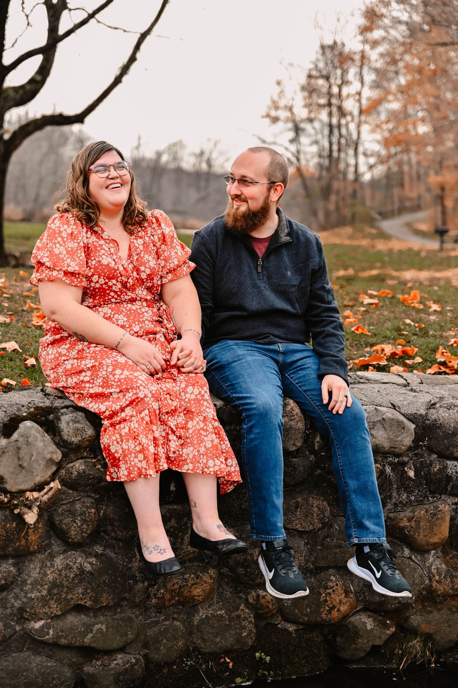 Happy couple sitting on stone wall during autumn engagement photoshoot.
