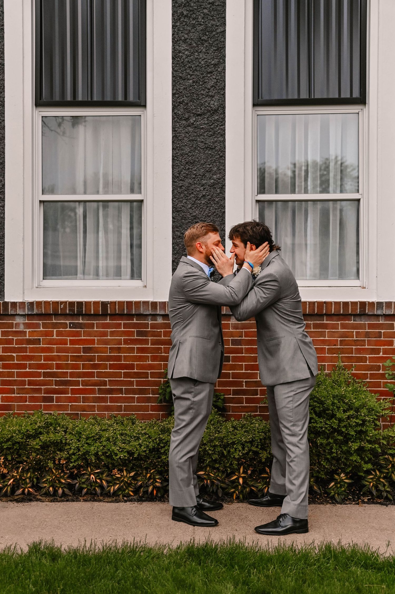 Wedding party members in gray suits lean forward in a synchronized pose against a brick wall.
