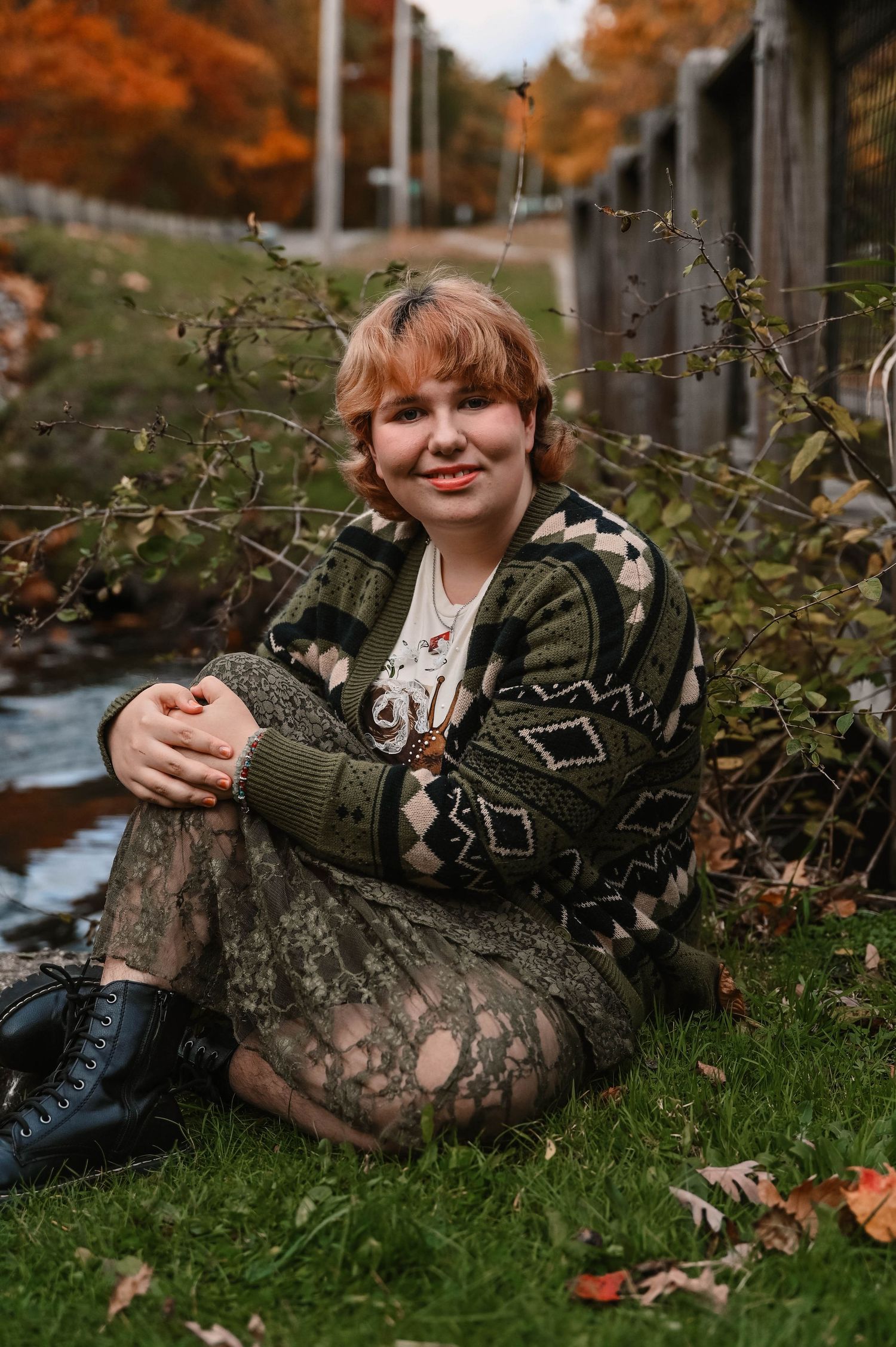 Person in patterned sweater relaxes on grass near autumn foliage.