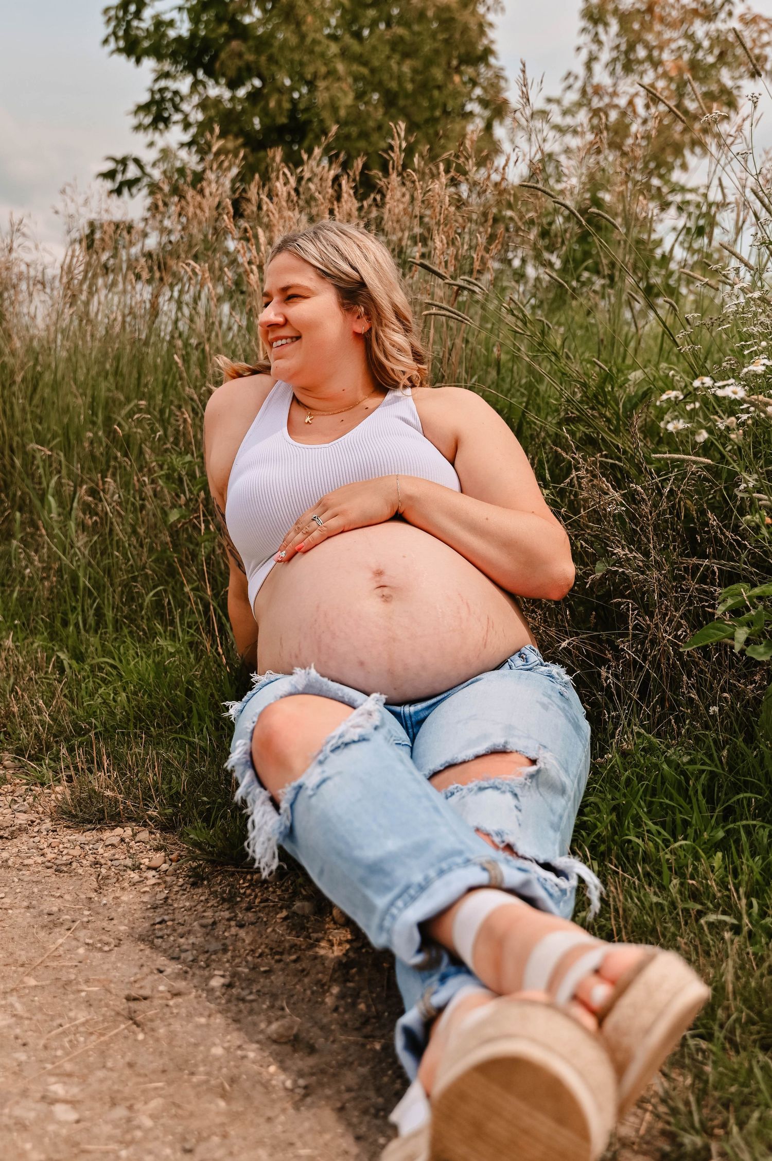 Casual maternity photos taken on a dirt path surrounded by tall grass and wildflowers.