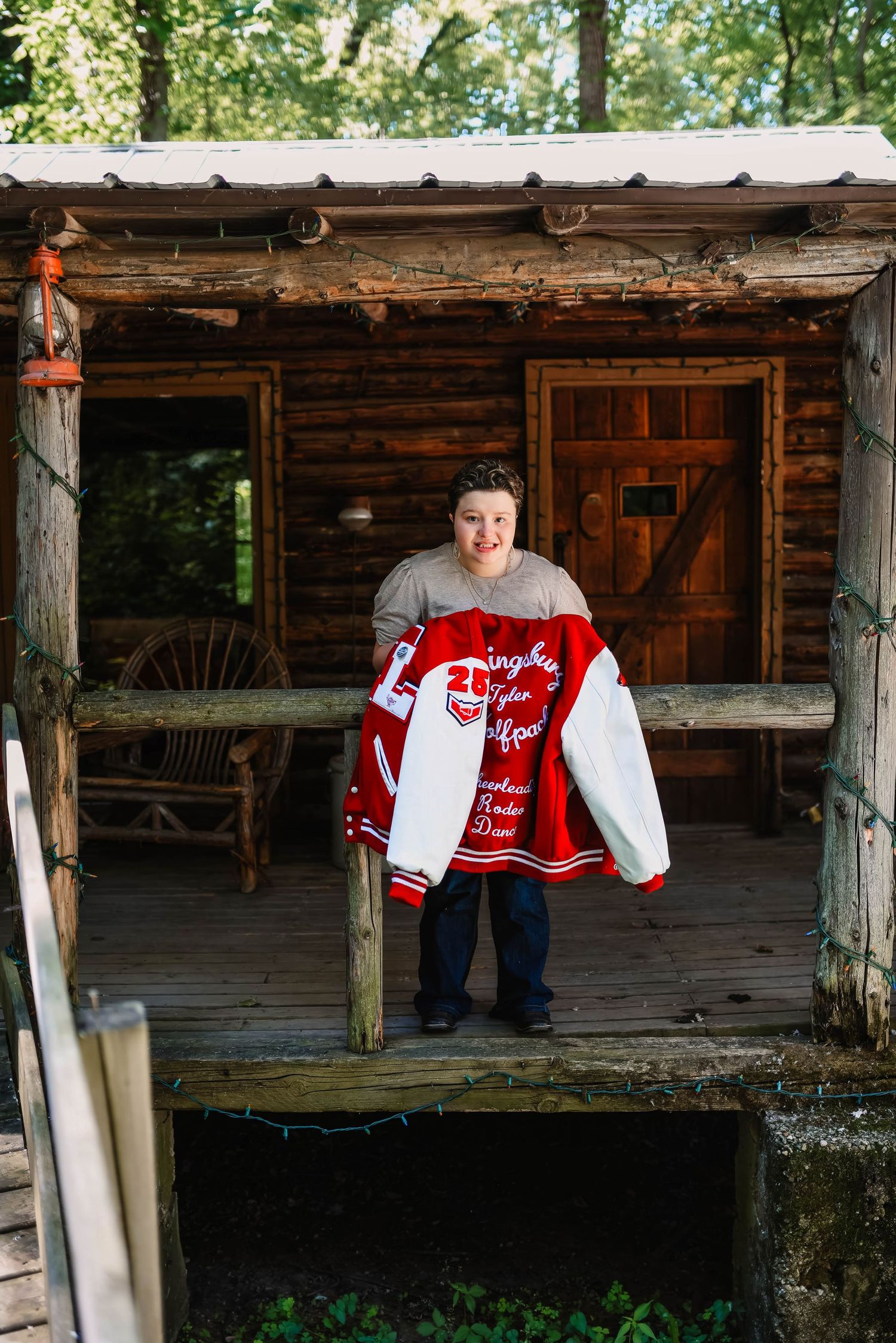 A person displays a red and white varsity jacket while standing on a rustic cabin porch.