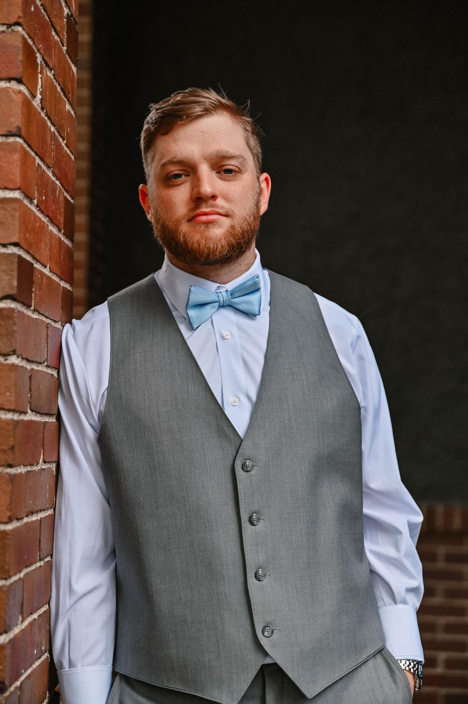 Groom in formal attire wearing a grey vest, light blue bowtie and white dress shirt poses against brick wall.