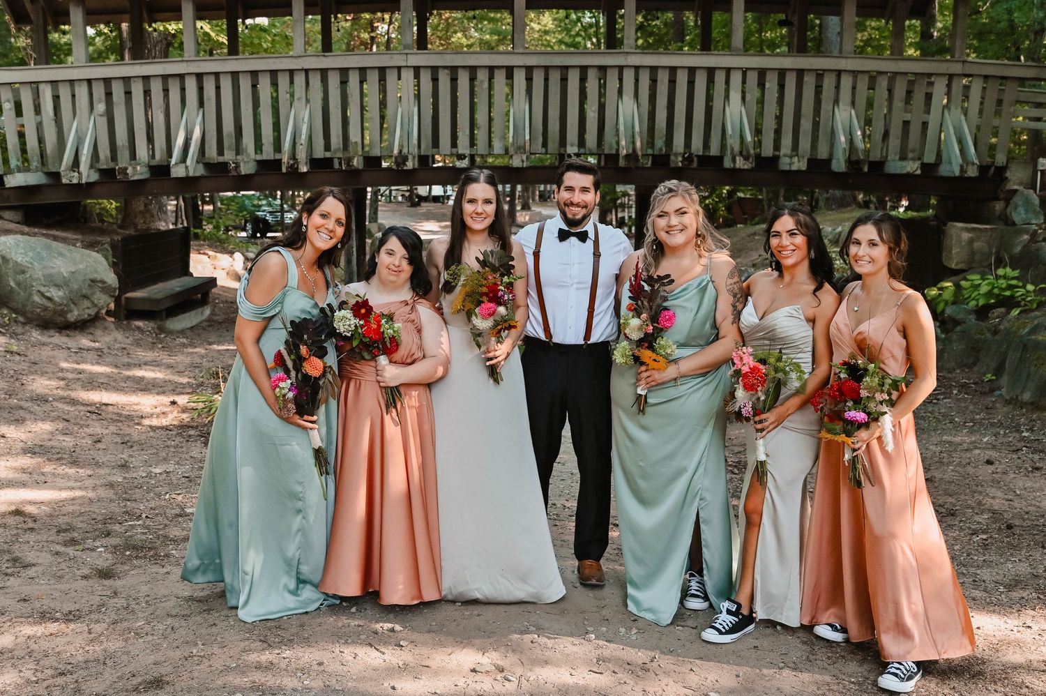 Wedding party in mint and peach dresses pose in front of rustic bridge.