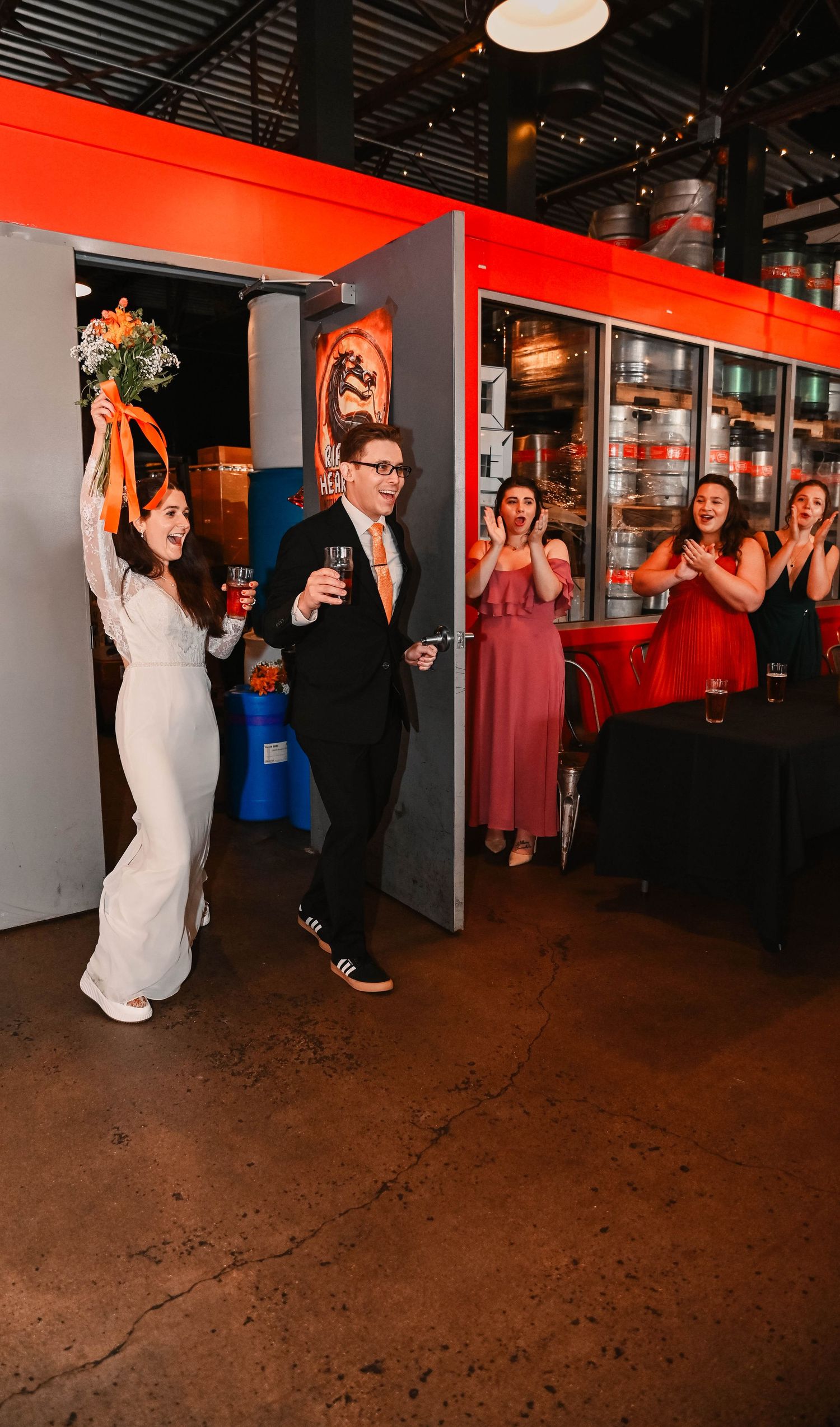 Wedding party performs choreographed dance moves in front of red wall at brewery venue.