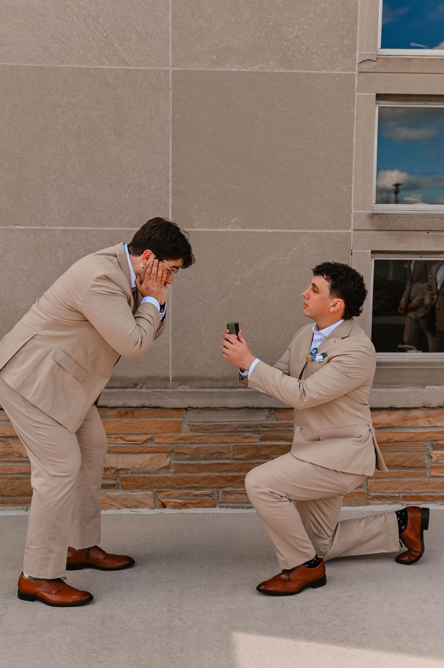 Two people in matching beige suits acting out a surprise proposal scene outdoors.