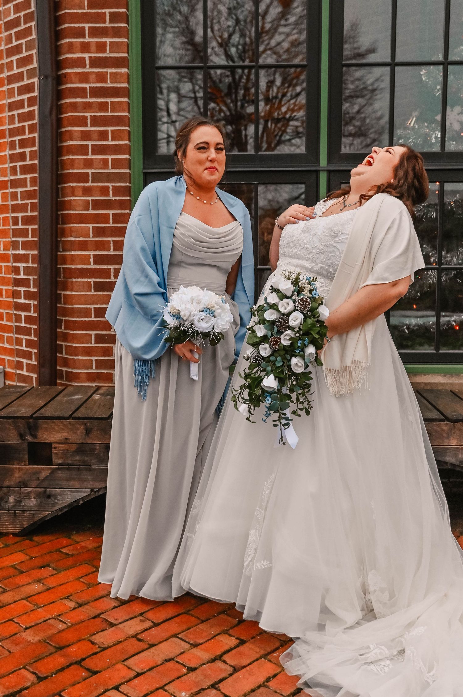 Bridesmaids in light gray dresses pose playfully on brick patio holding blue bouquets.