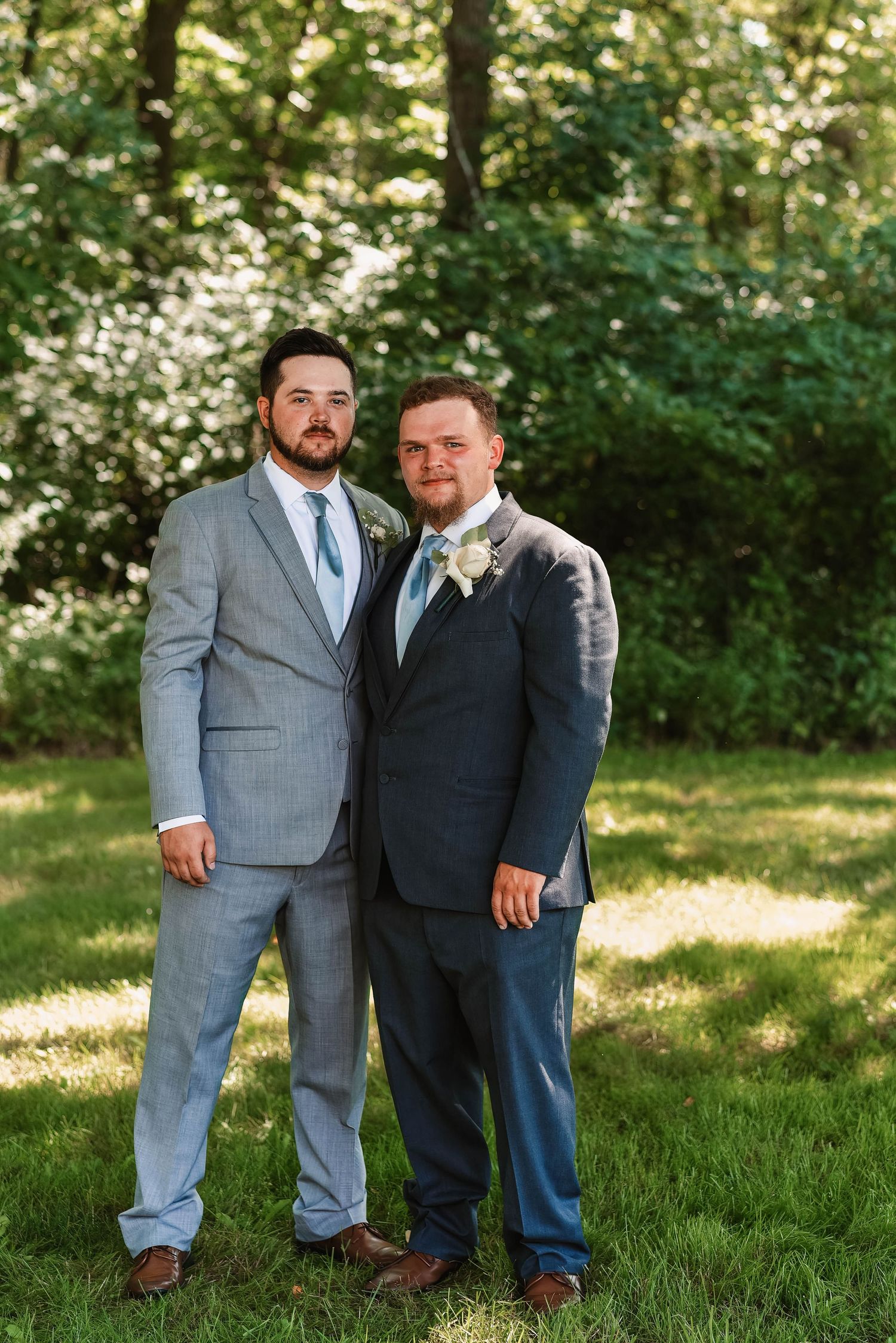 Two groomsmen in formal attire pose together in a sunny outdoor garden setting.