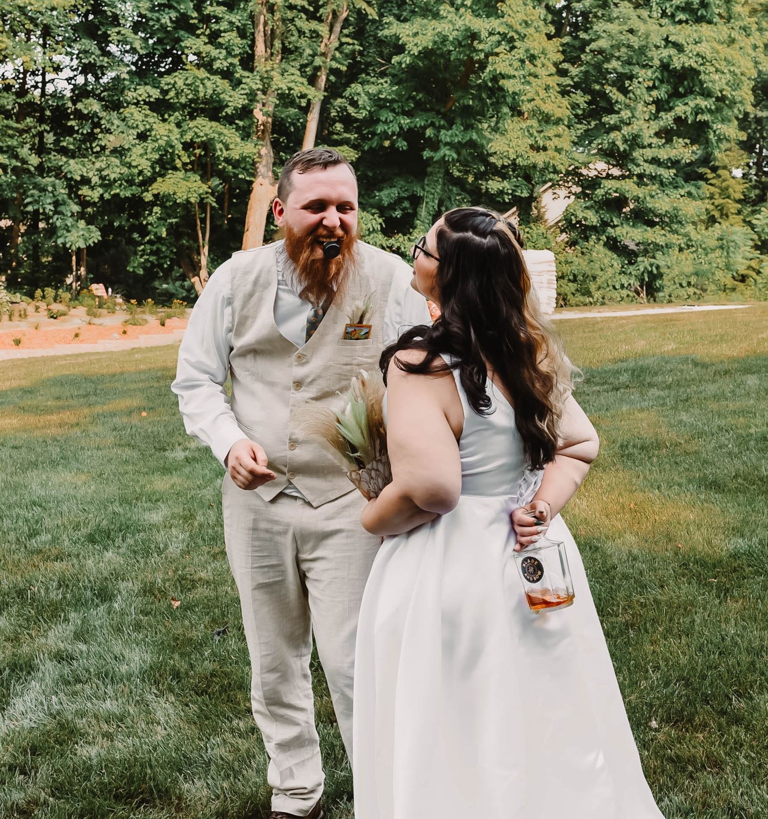 Couple in wedding attire walking together on grass with trees in background.
