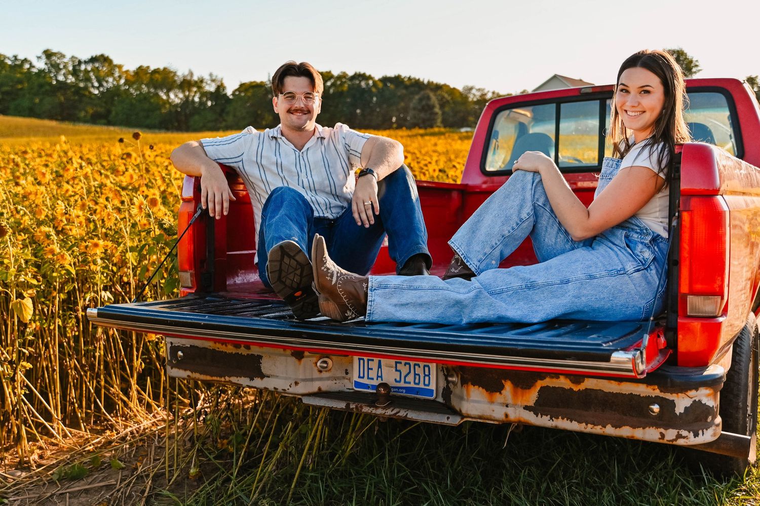 Couple relaxing in red pickup truck bed surrounded by sunflowers at sunset.