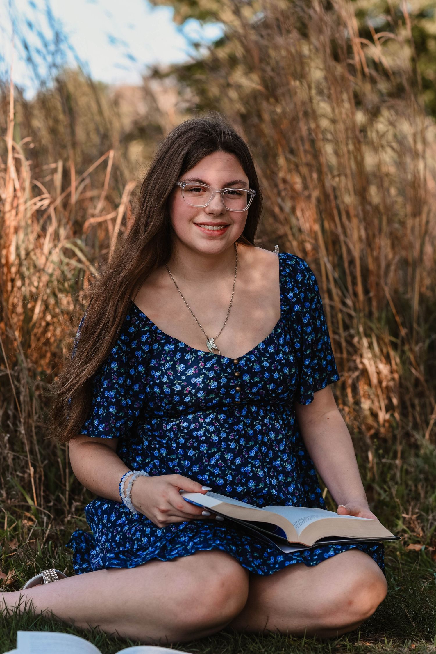 Reading outdoors in a blue floral dress among tall grass at sunset.