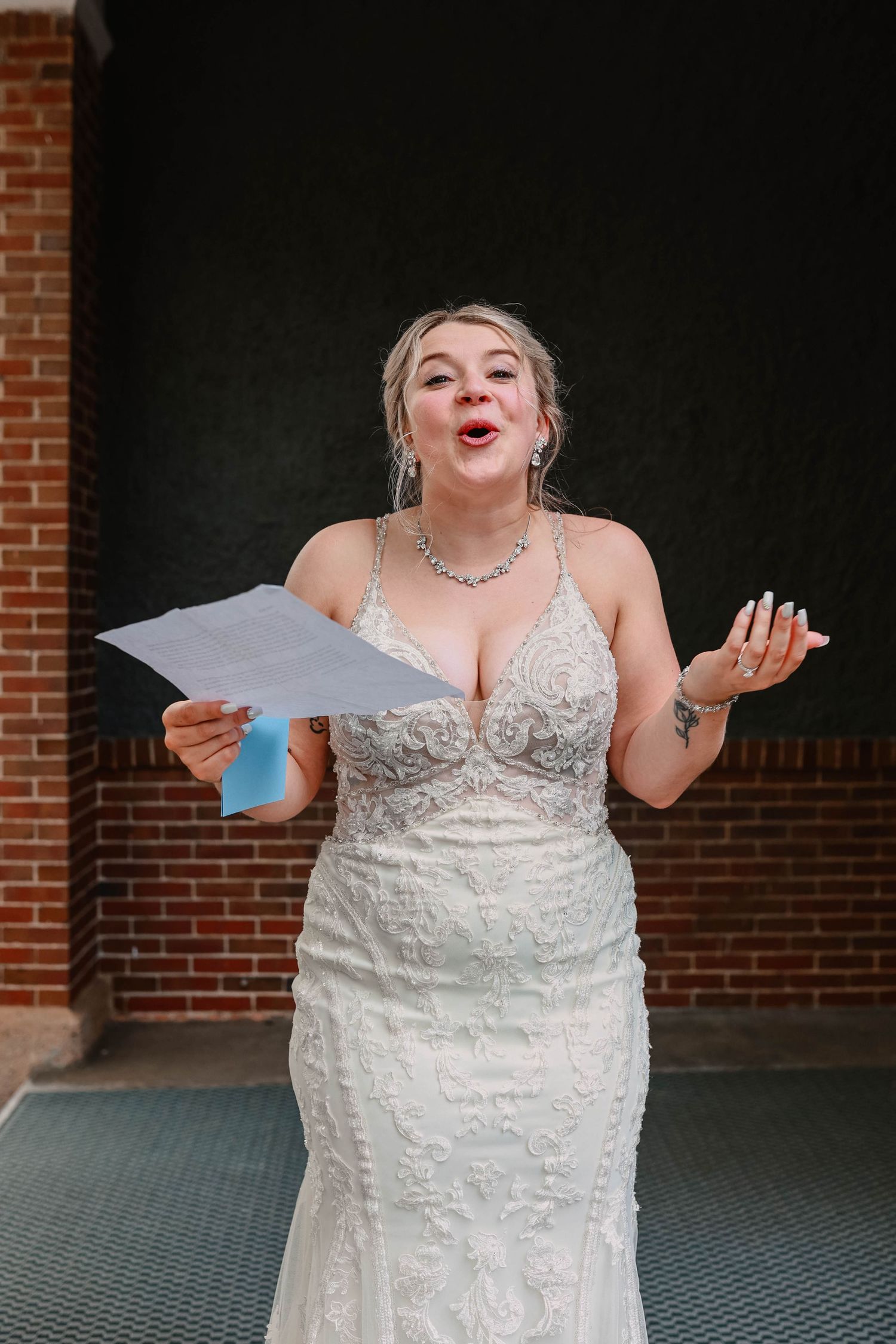 Bride in lace dress makes playful expression while holding papers outdoors.