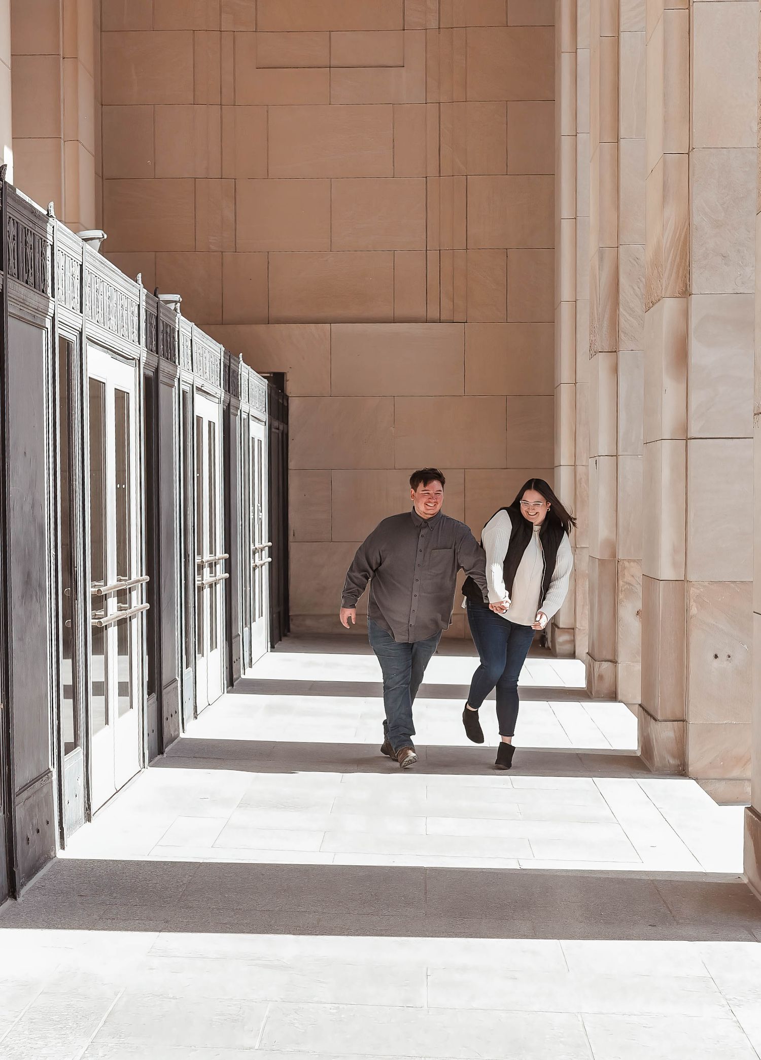 A couple walks through a modern colonnade with repeating architectural columns and shadows.