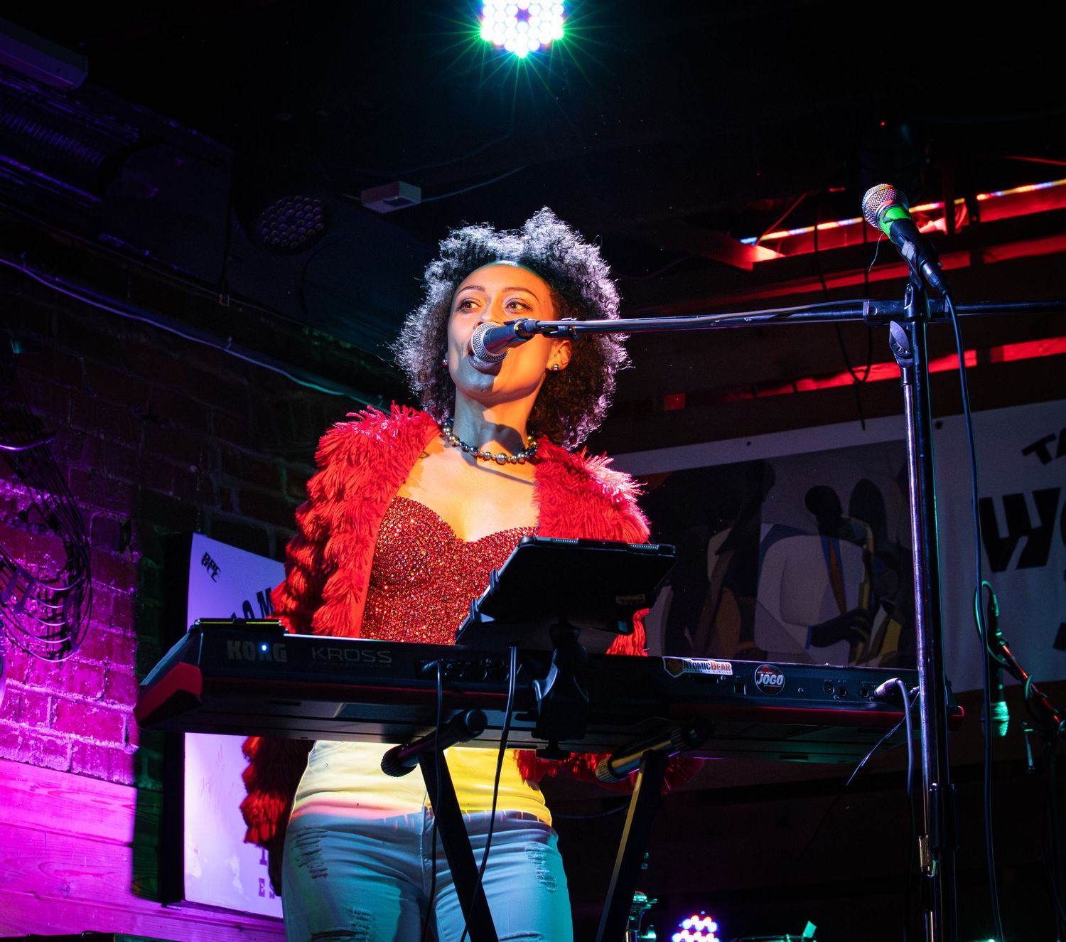 Performer in a red top sings passionately at a microphone on stage with colorful purple and red lighting.