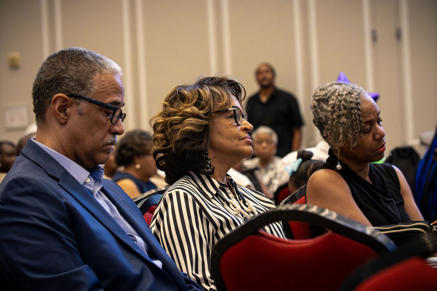Three attendees seated at a formal event, dressed in business attire, listening attentively in a crowded auditorium.