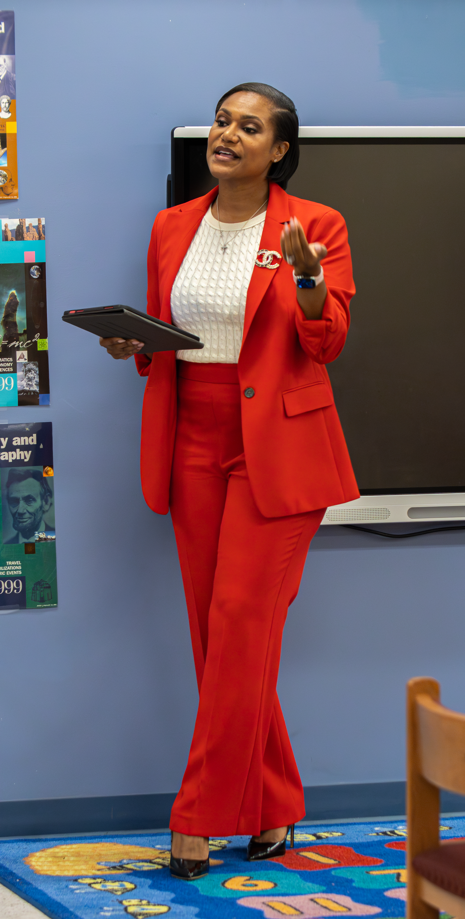 A woman in a red suit holds a tablet while gesturing in a colorful classroom near a smartboard.