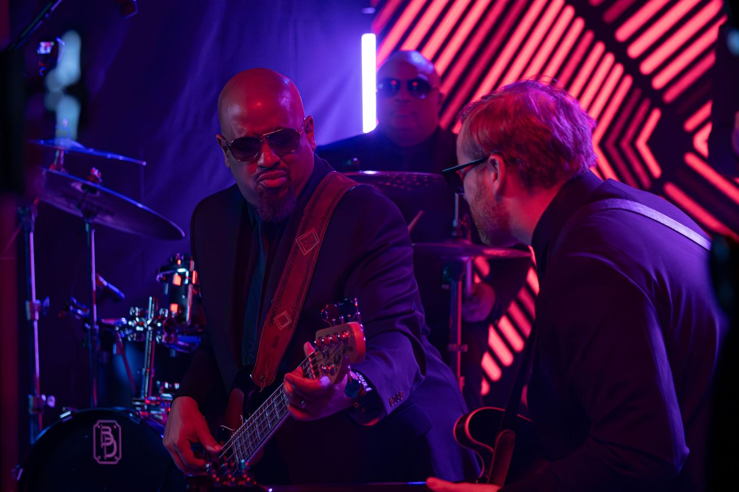 Band performs under vibrant red and purple stage lights with diagonal striped backdrop.