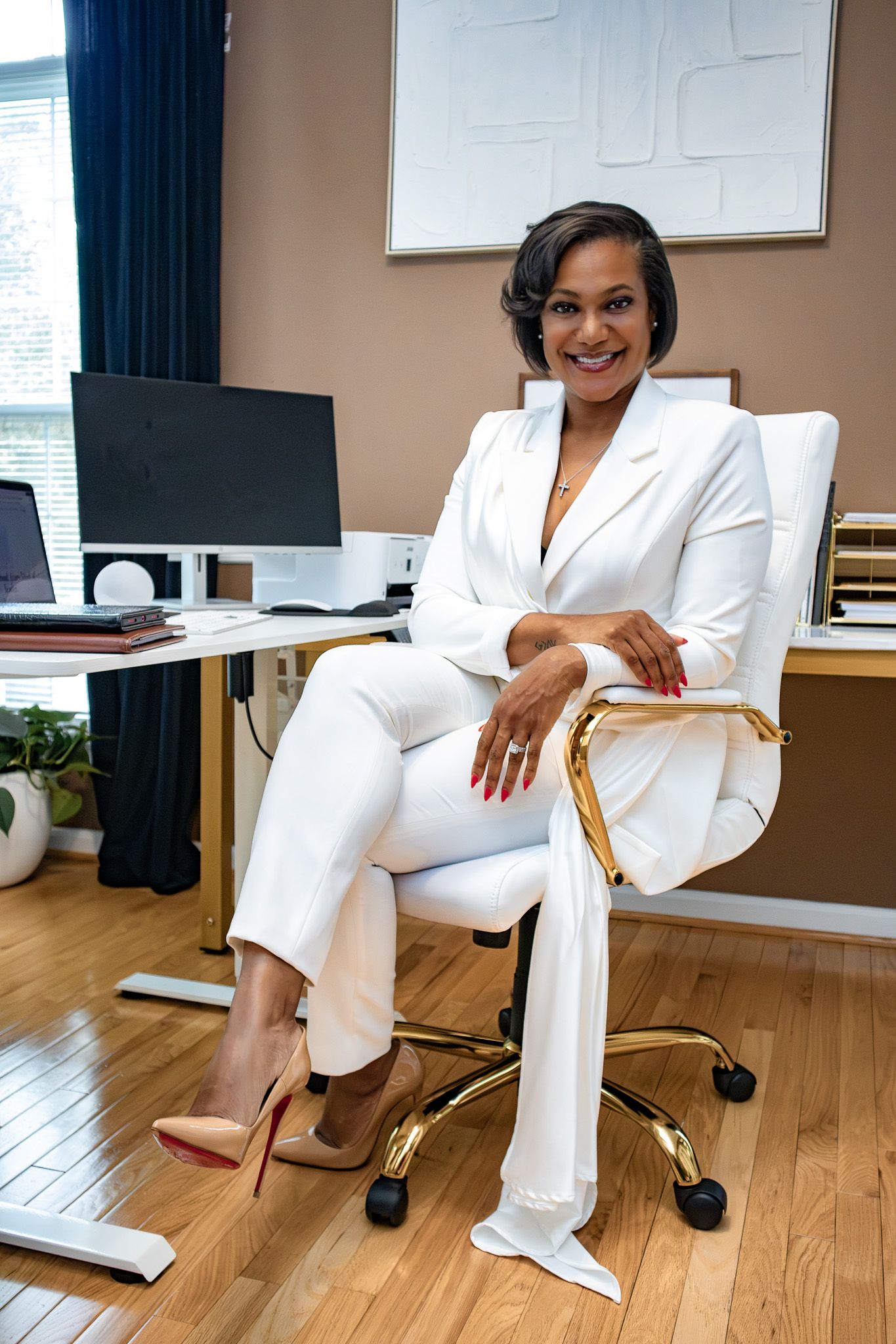 Professional Black woman in white suit seated in gold office chair, smiling confidently at desk.