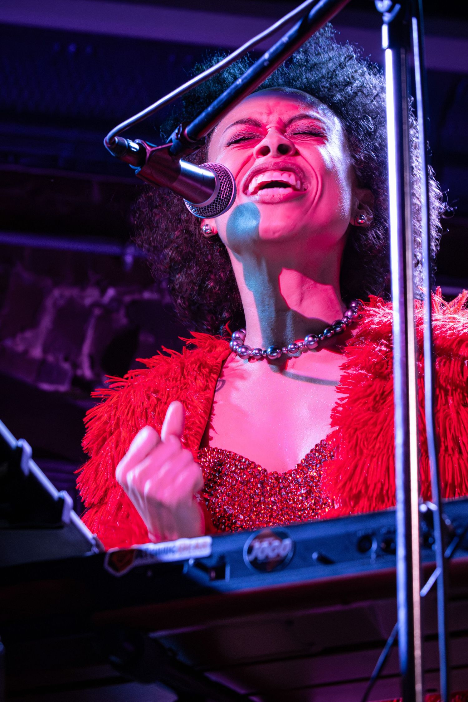 A performer in a red feathered outfit sings passionately at a microphone on stage under purple lighting.