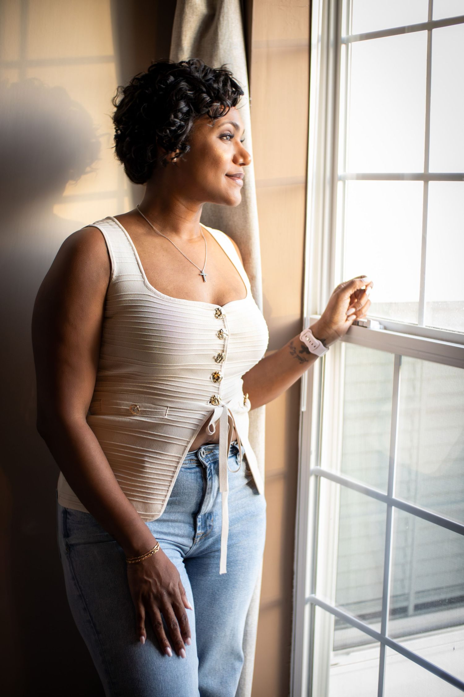 A Black woman in a white ribbed tank top and jeans gazes thoughtfully out a sunlit window.