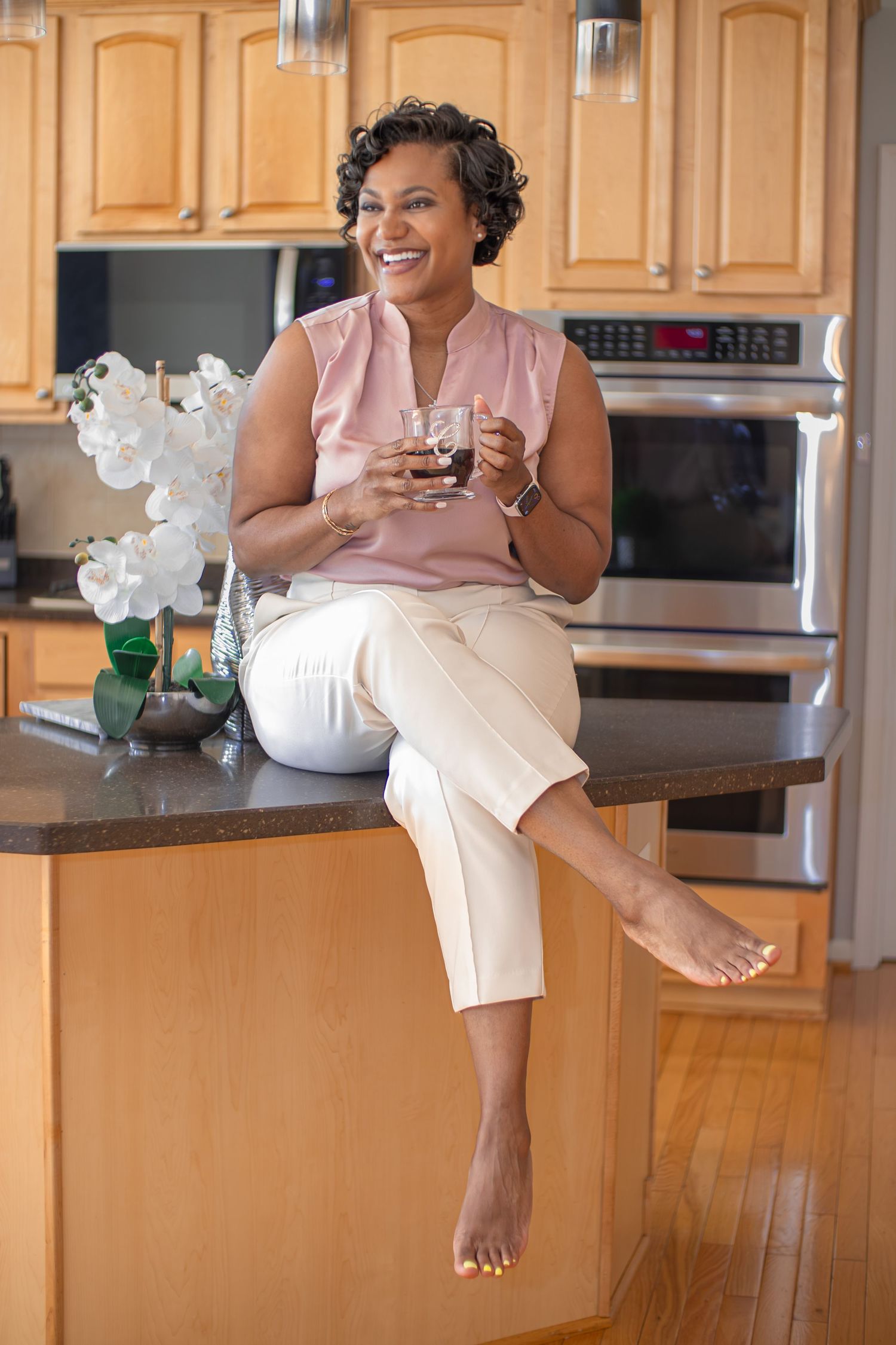 A smiling woman sits on a kitchen counter holding a glass of wine, wearing a pink blouse and white pants.