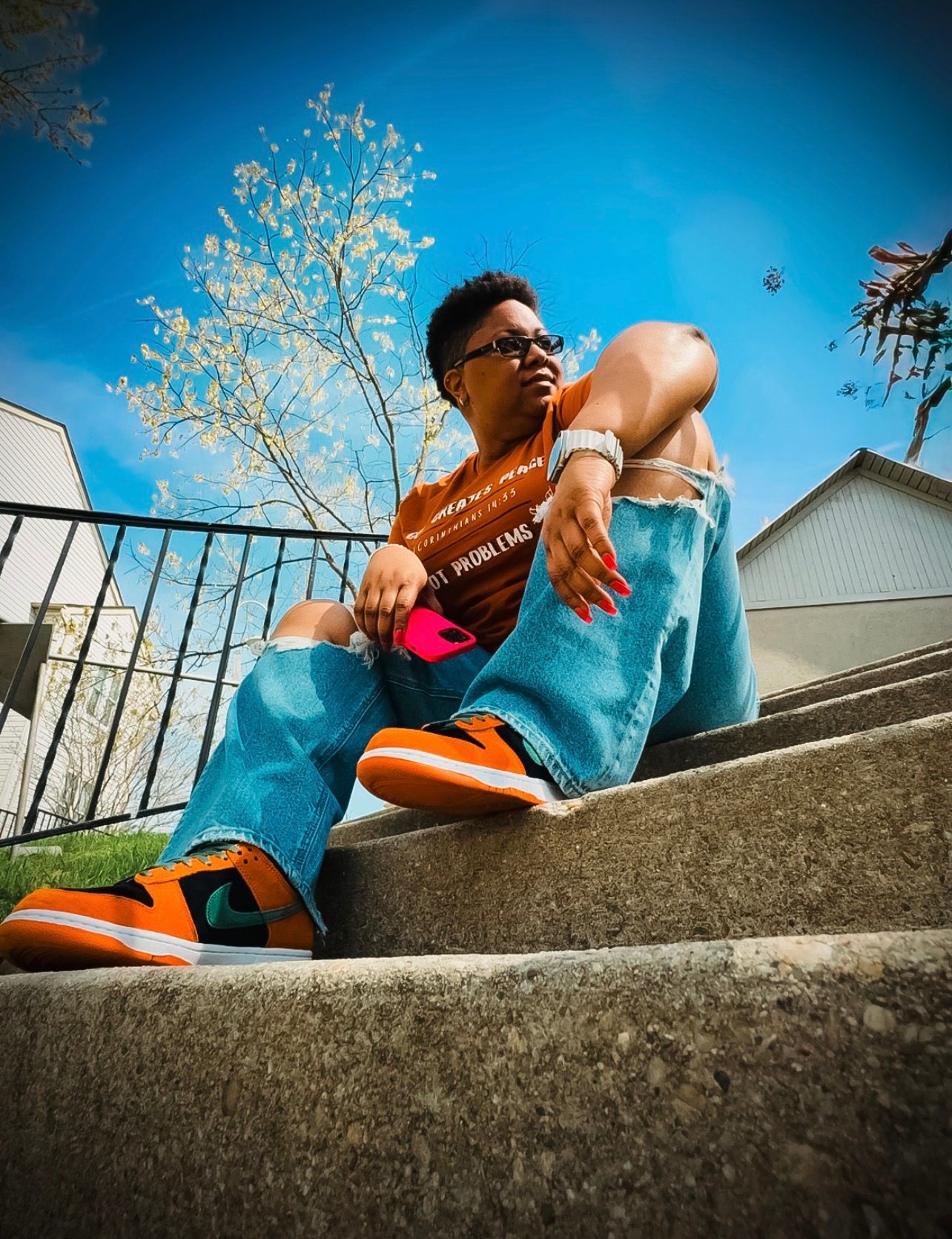 Young person sitting on concrete steps wearing orange sneakers, blue jeans, and an orange shirt under a blue sky.