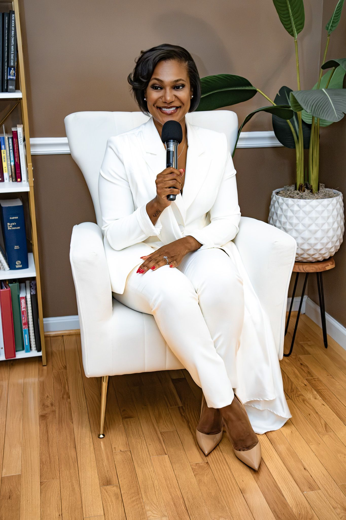 Confident Black woman in white suit holding microphone, seated in white chair with bookshelf backdrop.