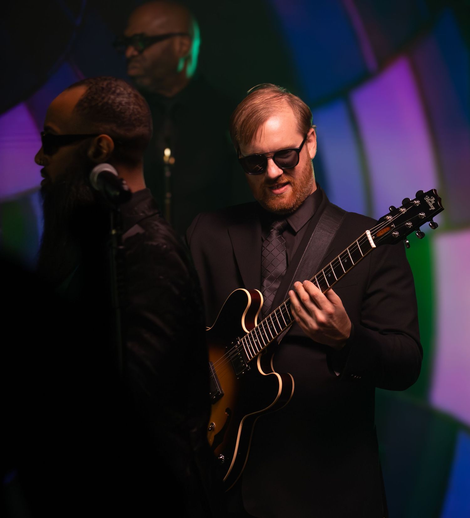 Guitar players perform on a dimly lit stage with moody blue and purple lighting.