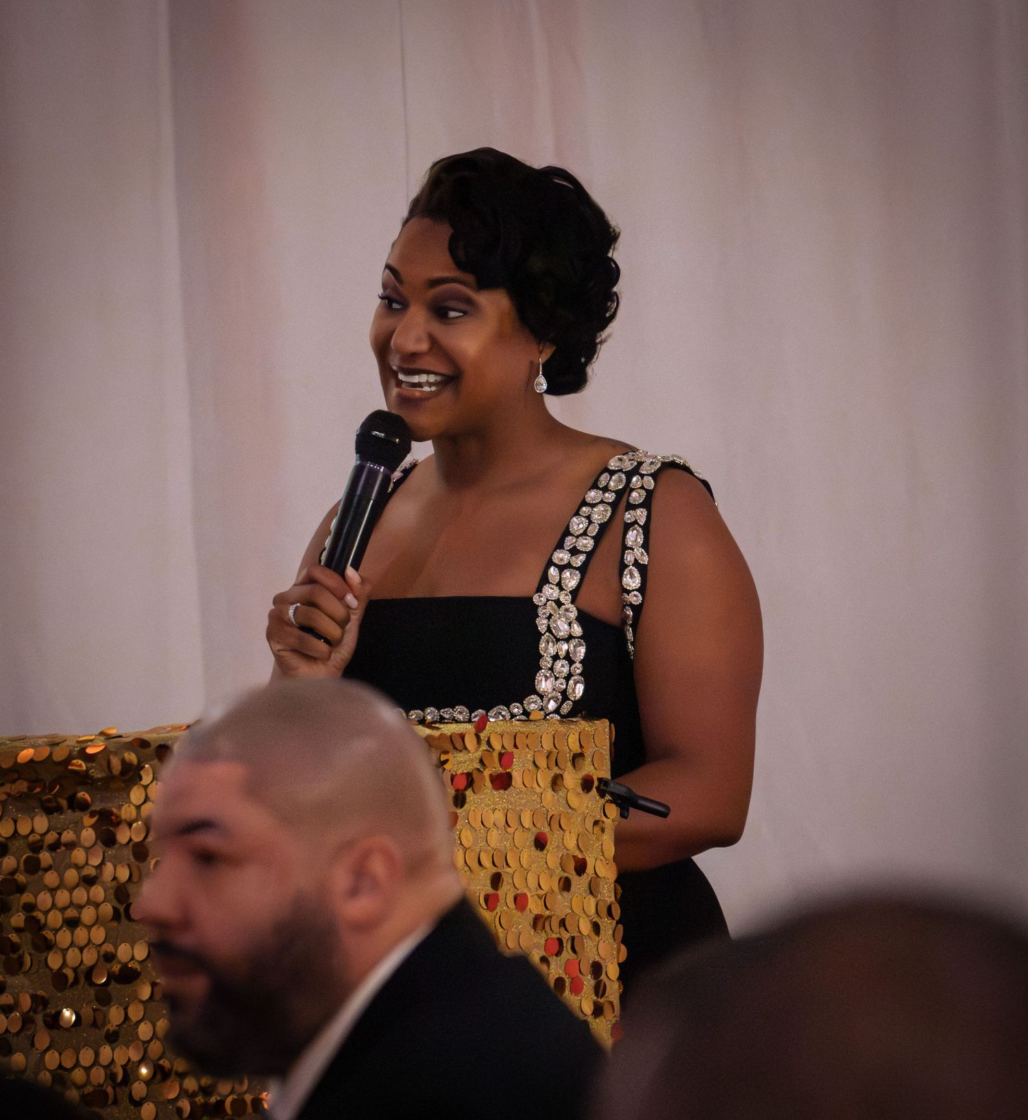 A speaker holds a microphone and gestures expressively during a presentation in front of a golden patterned backdrop.