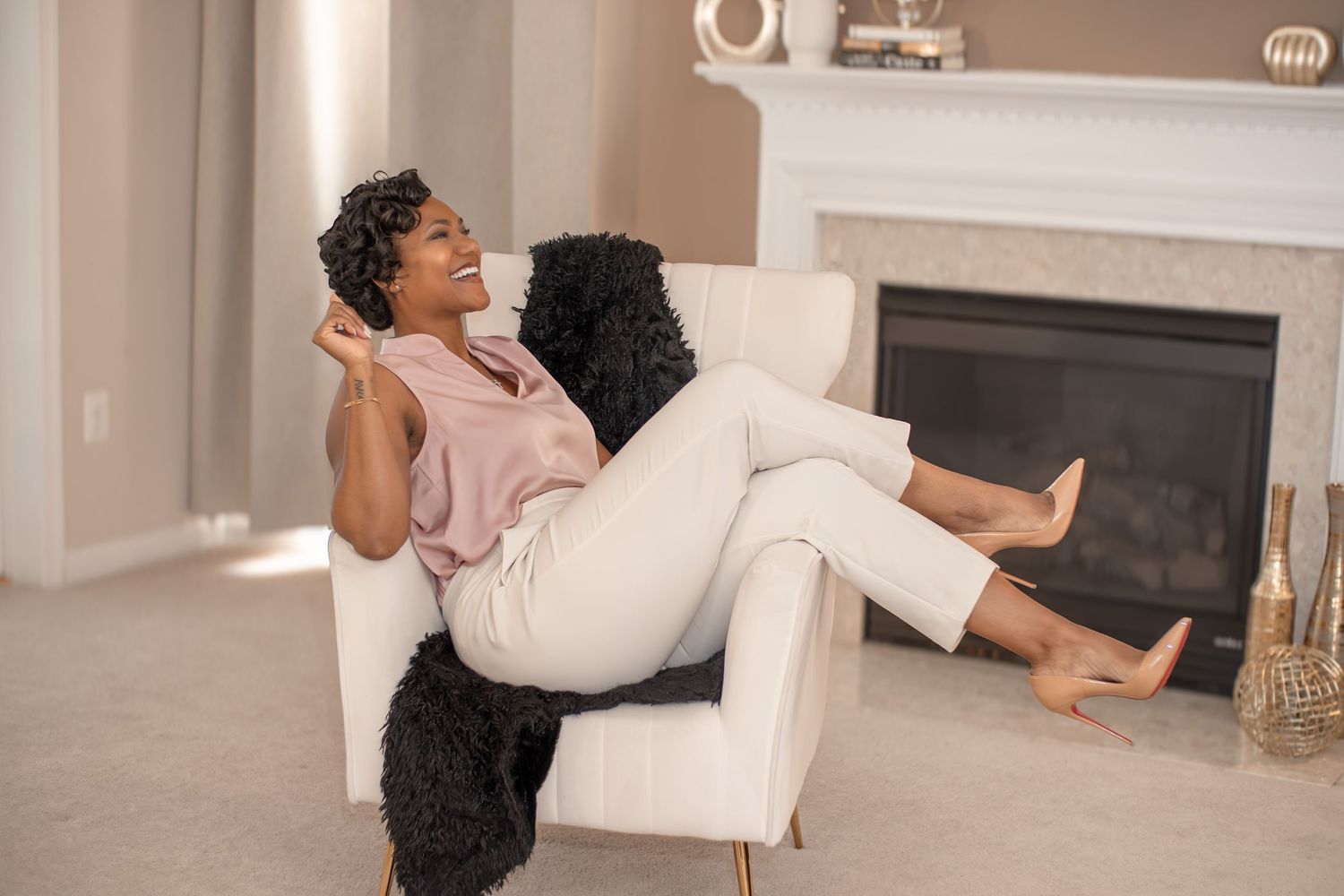 A laughing Black woman relaxes in a white armchair with a black dog beside her near a fireplace.