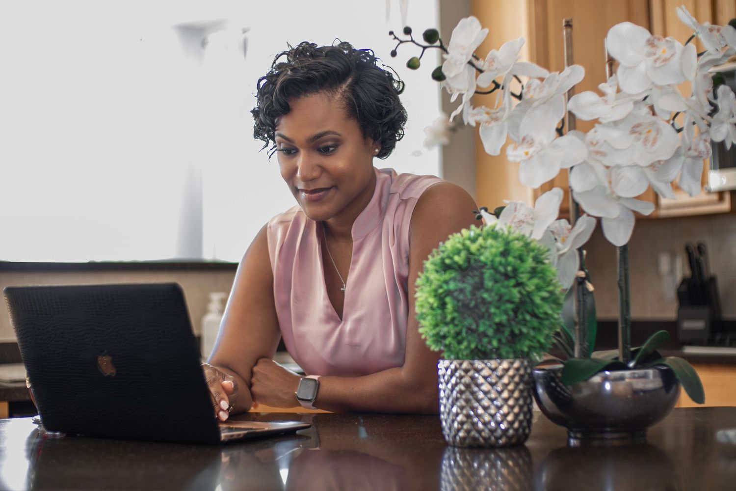 A woman working on a laptop at a wooden desk with potted plants beside her in a bright home office.