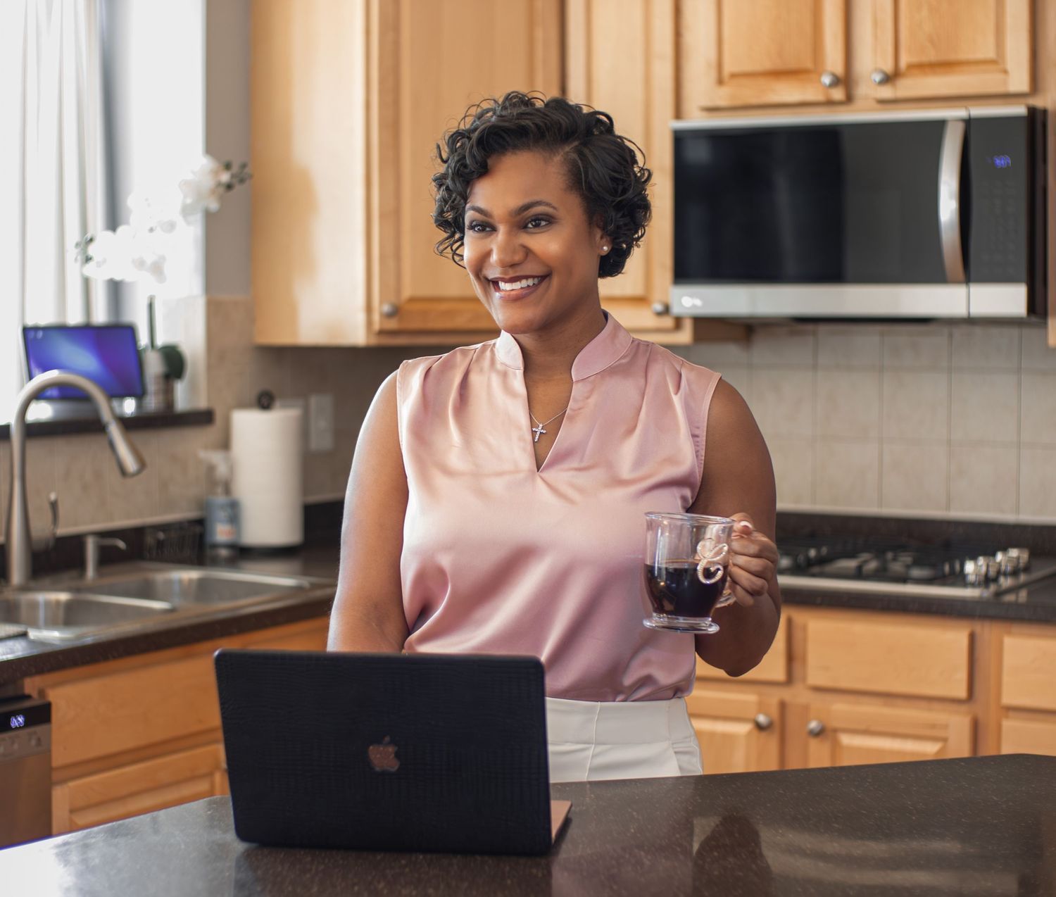 Woman smiling in kitchen holding coffee mug while working on laptop computer at home.