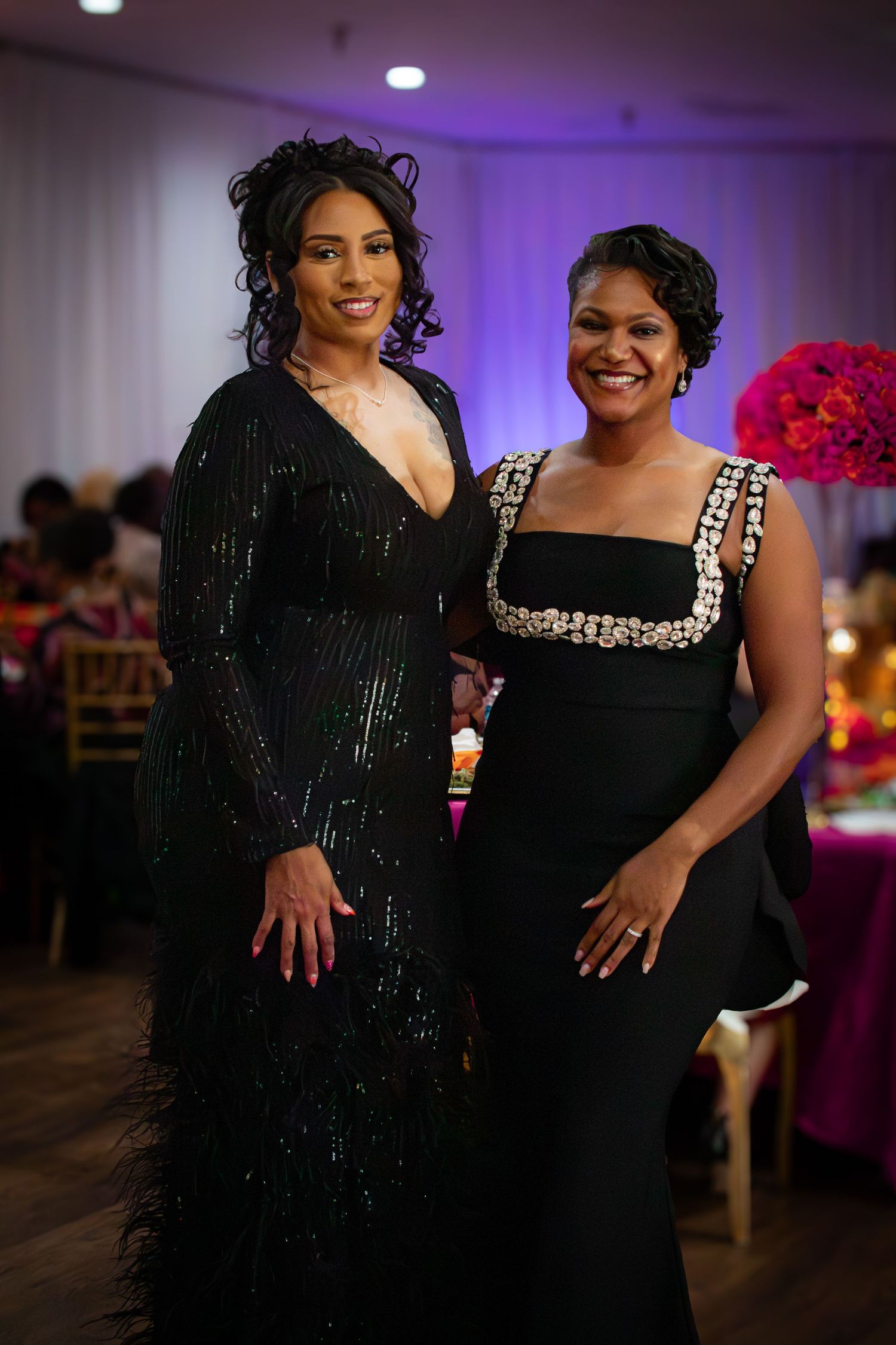 Two people wearing black evening attire pose together at a formal event with purple lighting in the background.