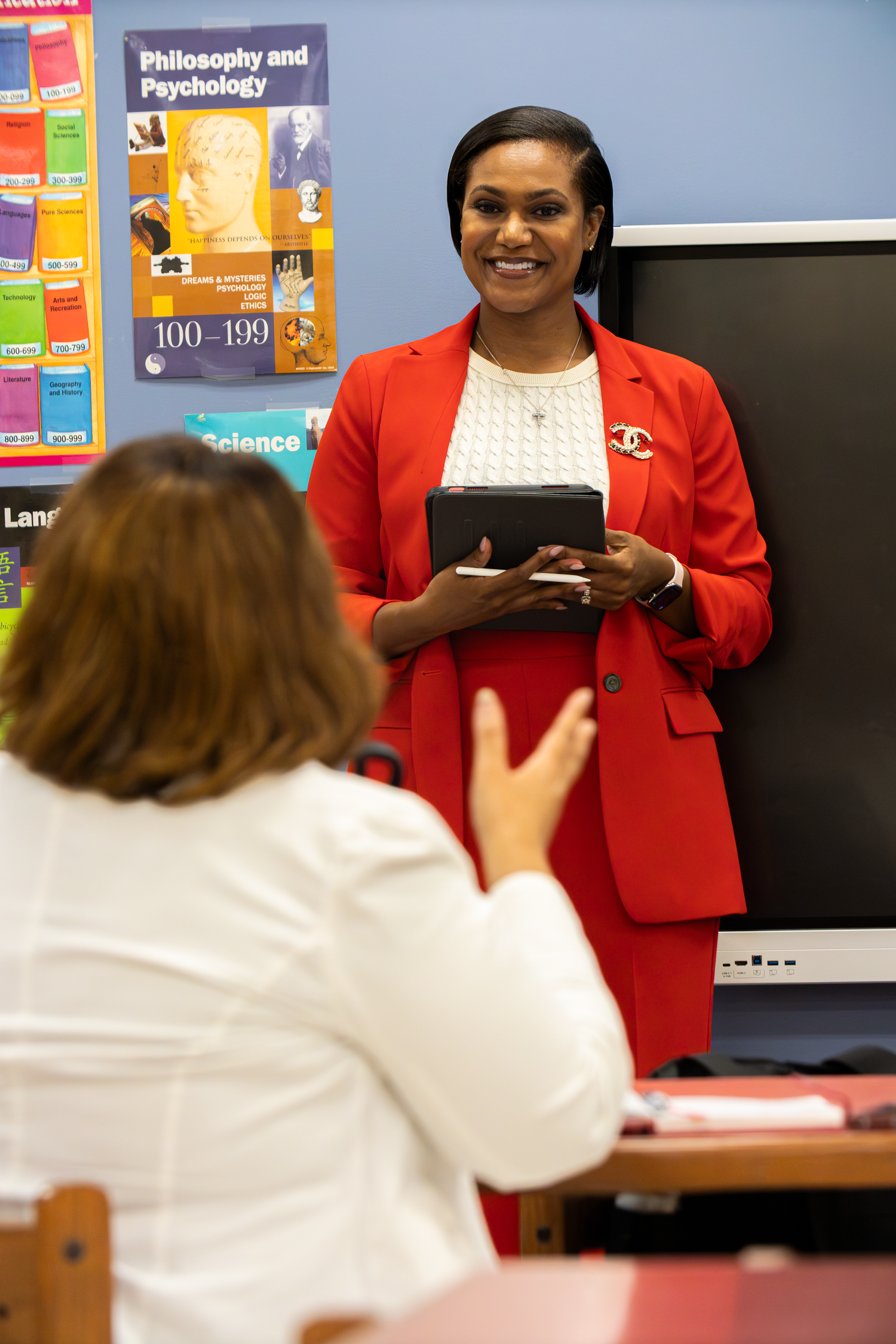 Woman in red suit holds tablet while smiling at student in colorful classroom setting.