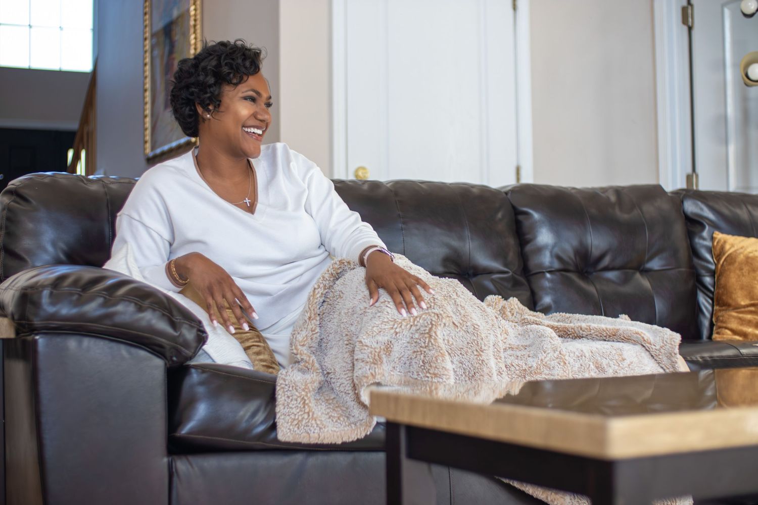 Happy woman relaxing on dark leather sofa with a decorative blanket in a bright living room.