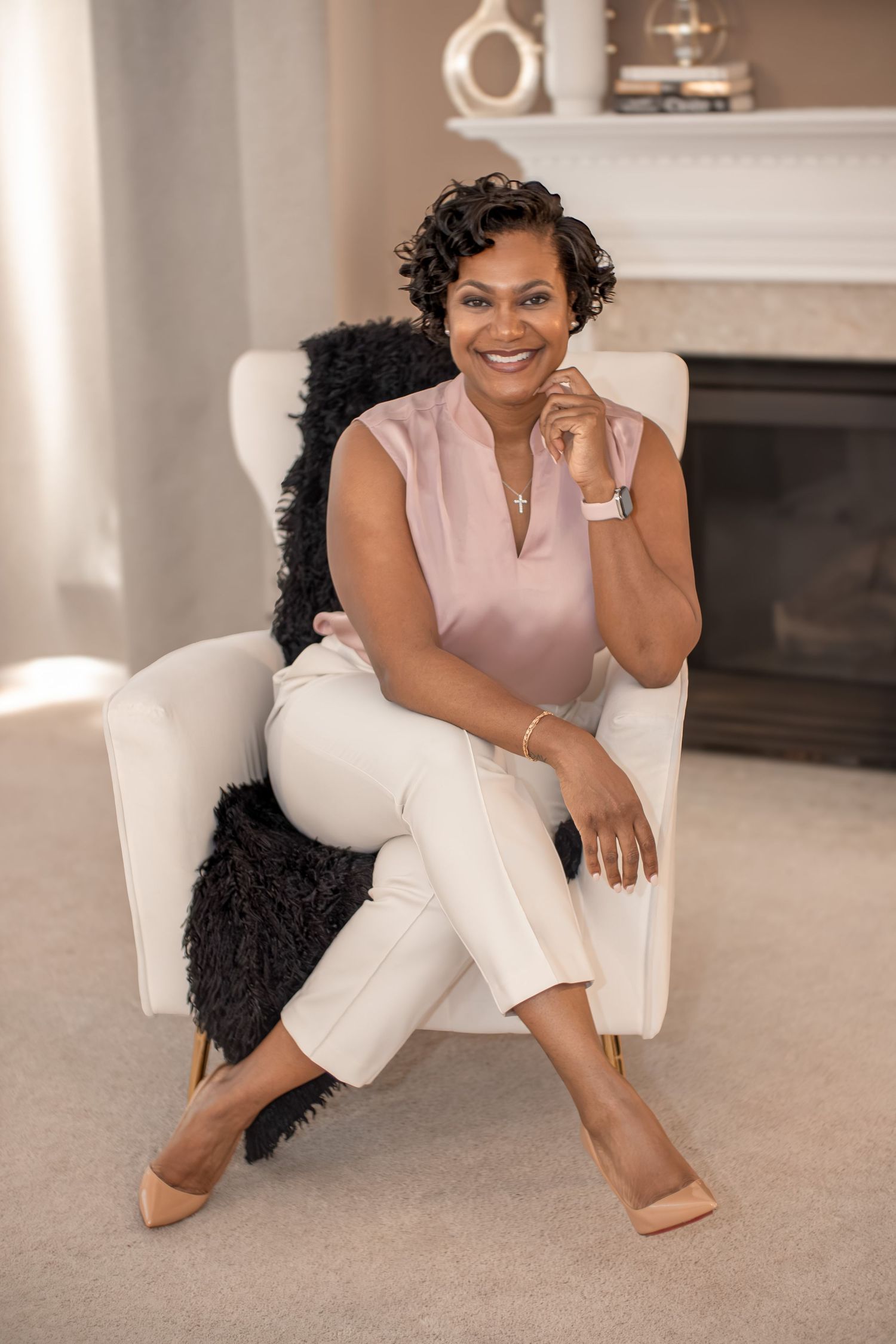 Smiling woman in pink blouse and white pants seated in white armchair near fireplace.