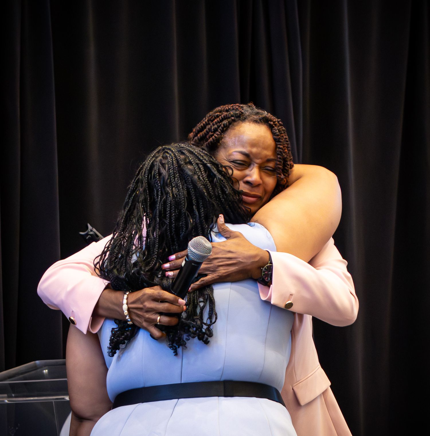 Two women sharing a warm embrace on stage, one holding a microphone, dressed in light-colored blazers at a formal event.